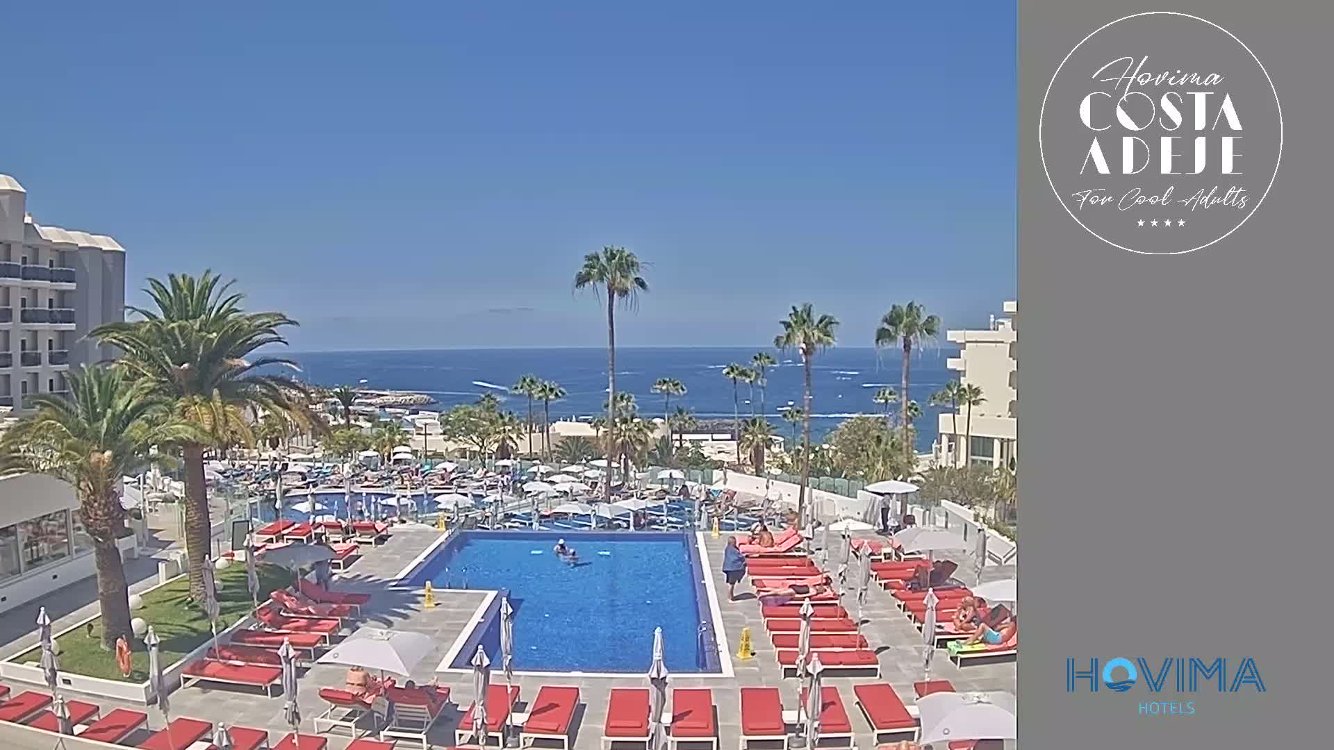 A resort pool area with red lounge chairs and umbrellas surrounds a swimming pool, all overlooking a calm ocean under a clear blue sky.