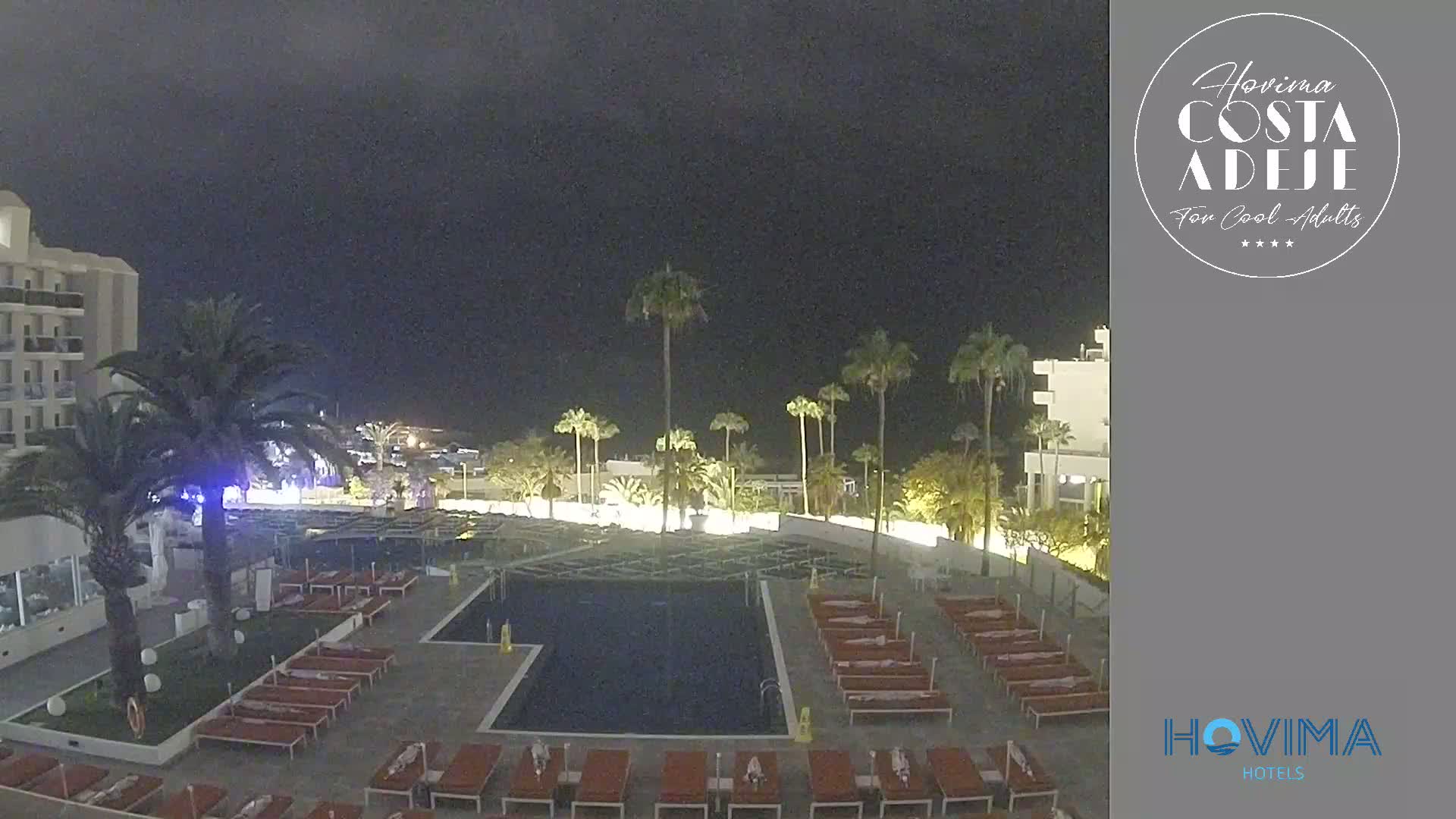 An outdoor nighttime scene shows a resort pool surrounded by lounge chairs and palm trees, adjacent to a multi-story building under a dark sky.