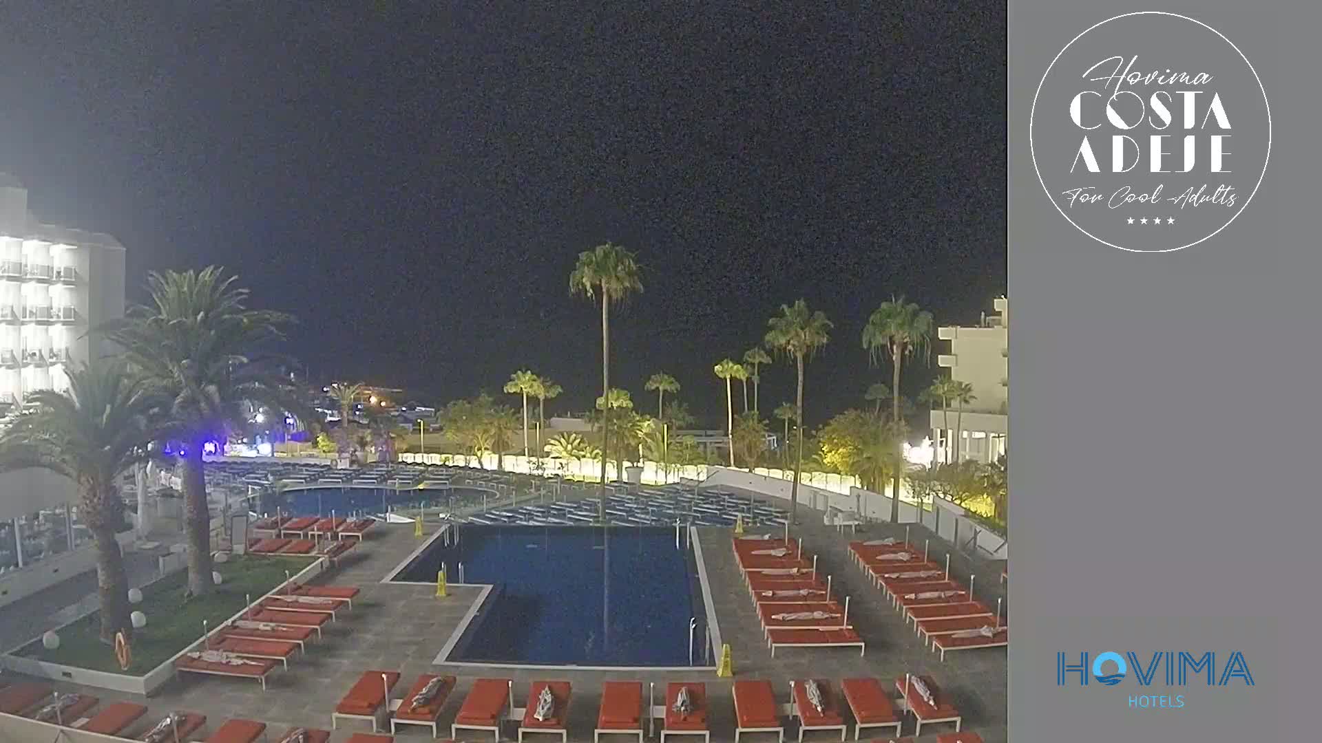 A nighttime view of a resort pool area with palm trees, lounge chairs, and a building under a clear, dark sky.