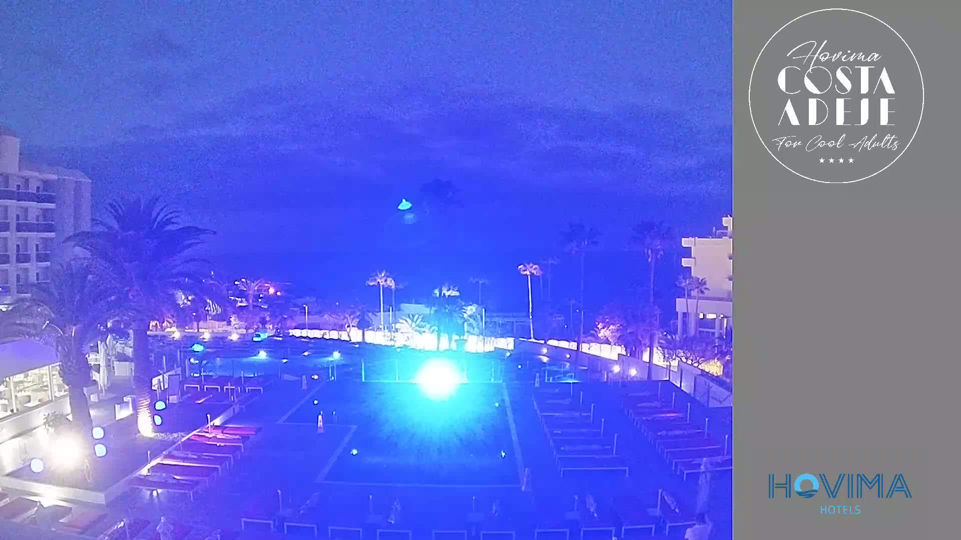 A nighttime view of a resort pool area with palm trees and hotel buildings under a dark blue sky.
