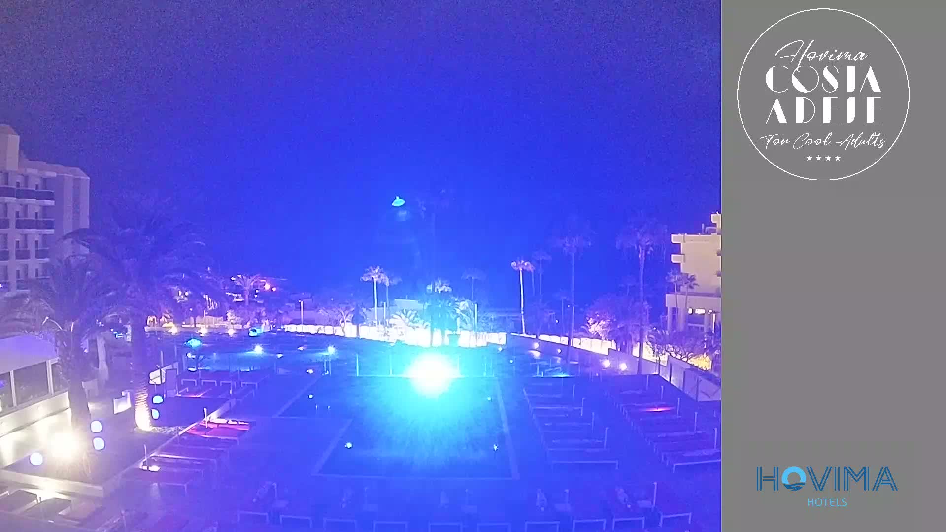 A brightly lit pool area at night, surrounded by palm trees and hotel buildings, under a dark blue sky.