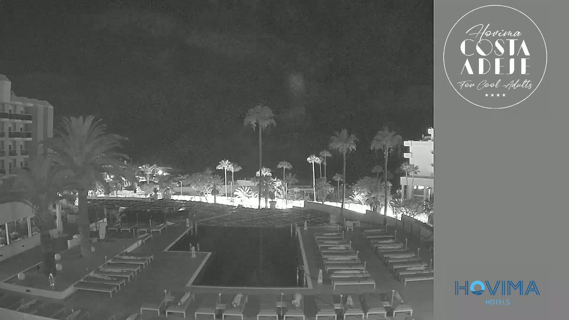 A nighttime, low-light view of a resort pool area with palm trees and lounge chairs surrounding a dark pool, next to a multi-story building.