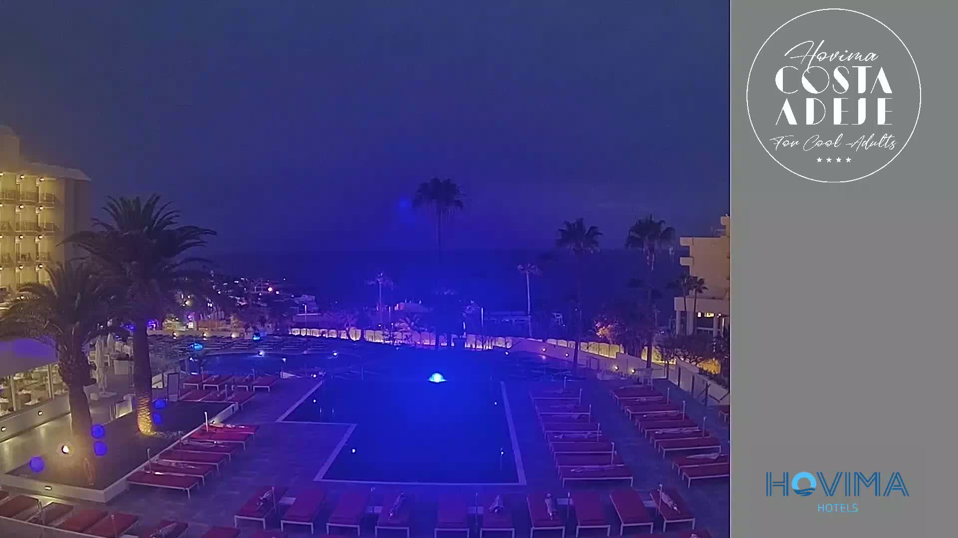 A nighttime view of a hotel pool area with palm trees, illuminated in blue light, under a dark blue sky.