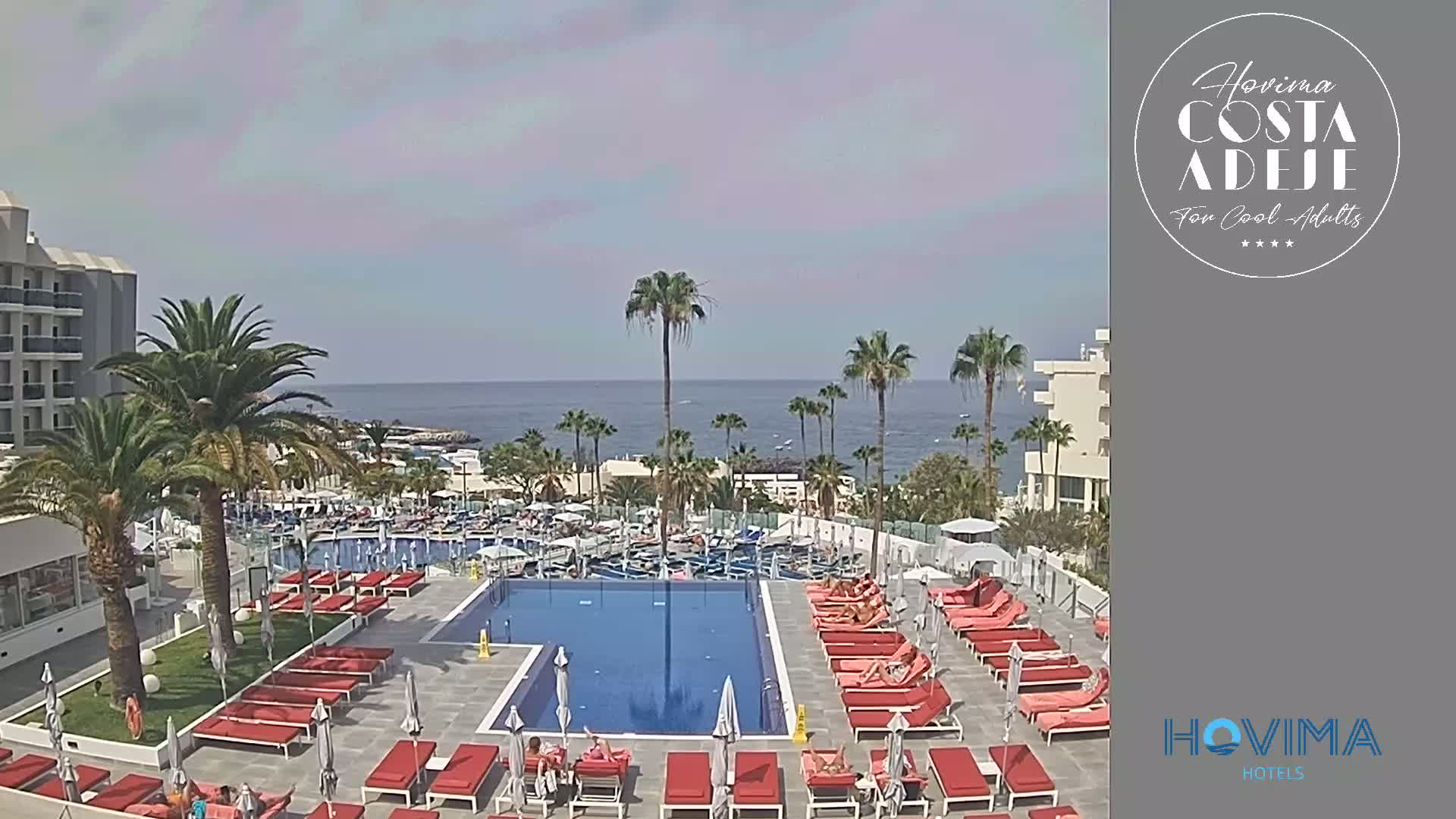 A resort pool area with red lounge chairs surrounds a large rectangular pool, overlooking a calm ocean under a mostly sunny sky.