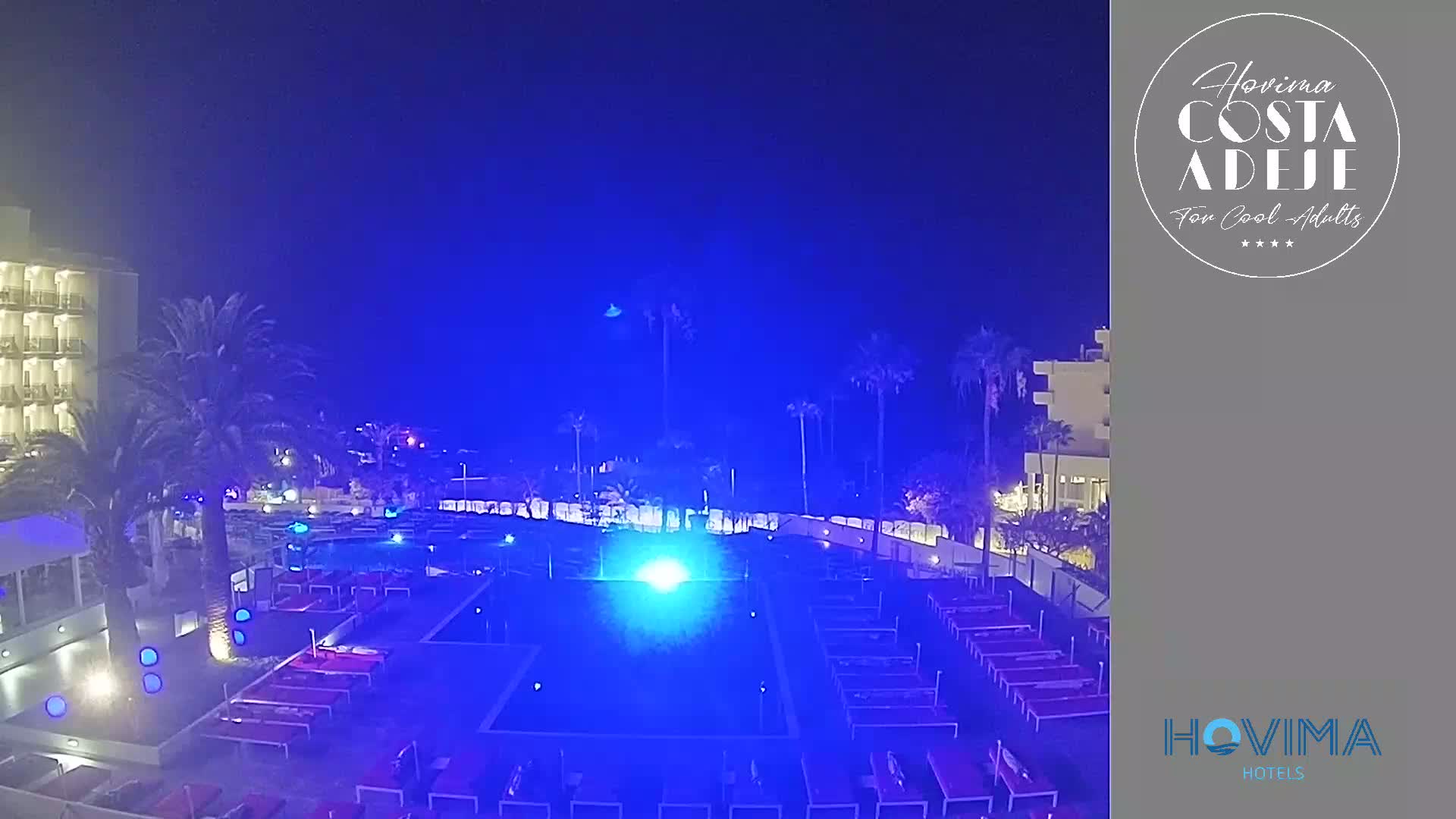A nighttime view of a resort pool area illuminated in blue light, with palm trees and a hotel building visible in the background under a clear, dark sky.