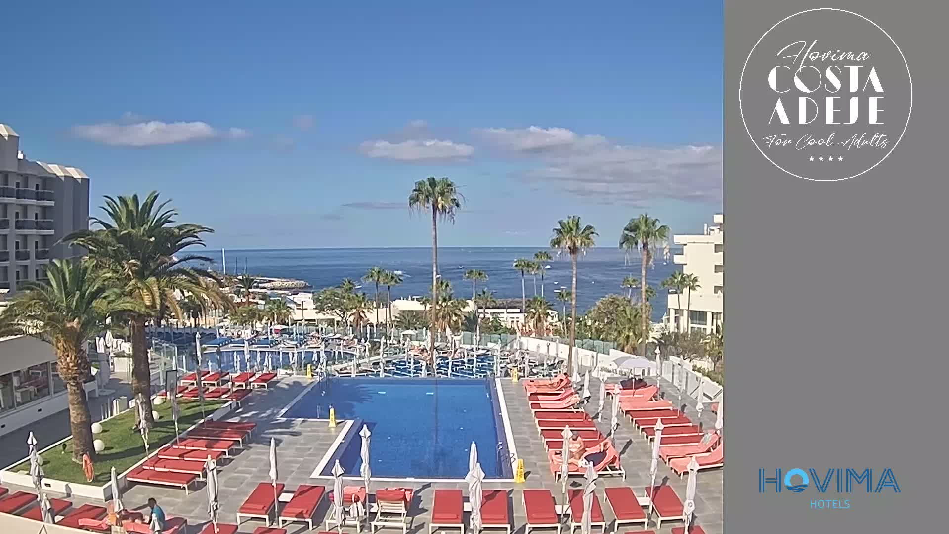 A resort pool area with numerous red lounge chairs surrounds a large, rectangular swimming pool, overlooking a calm ocean under a clear blue sky.