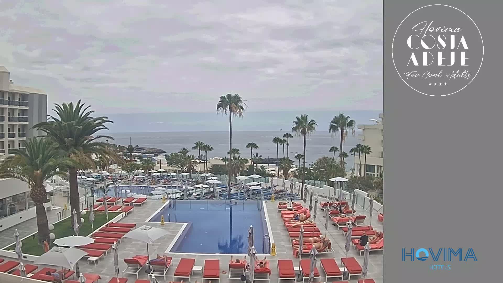 An outdoor pool area with red lounge chairs and umbrellas surrounds a rectangular pool, situated near a hotel and overlooking a calm ocean under an overcast sky.