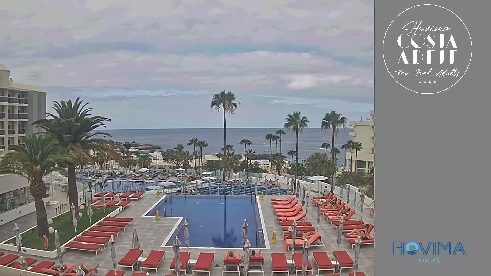 A resort pool area with red lounge chairs surrounds a swimming pool, overlooking a calm ocean under a cloudy sky.