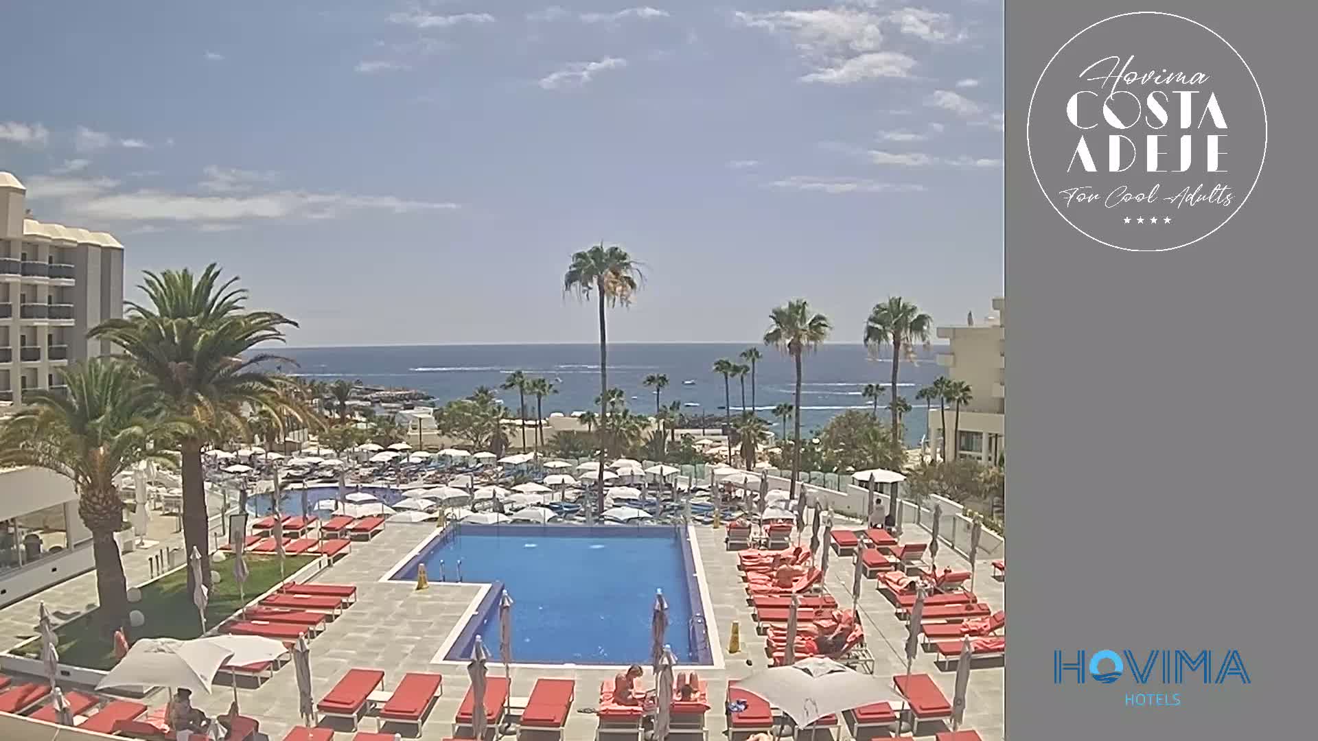 A resort pool area with red lounge chairs and umbrellas surrounds a large swimming pool, overlooking a calm ocean on a sunny day.