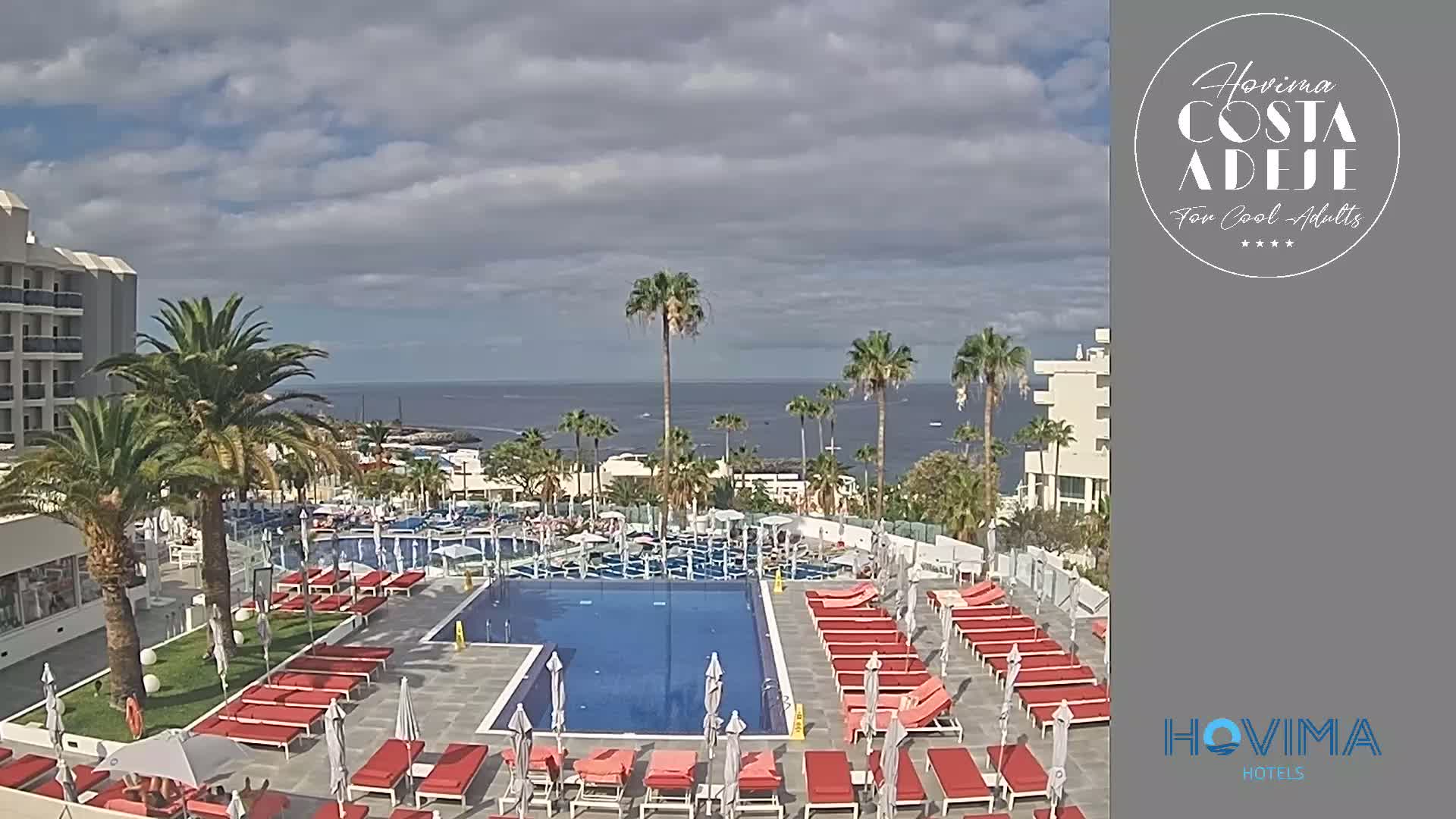 A resort pool area with red lounge chairs and umbrellas, palm trees, and a view of the ocean under a partly cloudy sky.