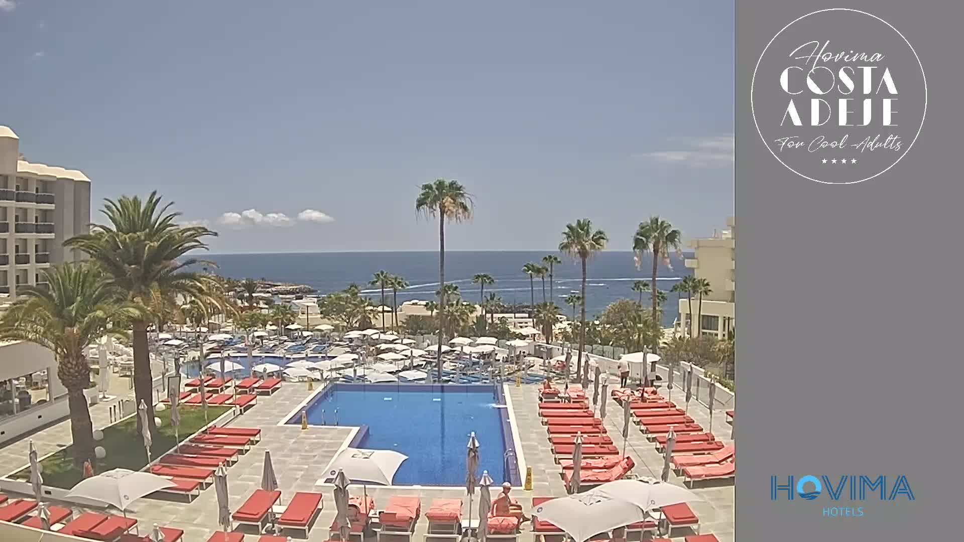 A resort pool area with red lounge chairs and umbrellas sits next to a building, overlooking a calm ocean on a sunny day.