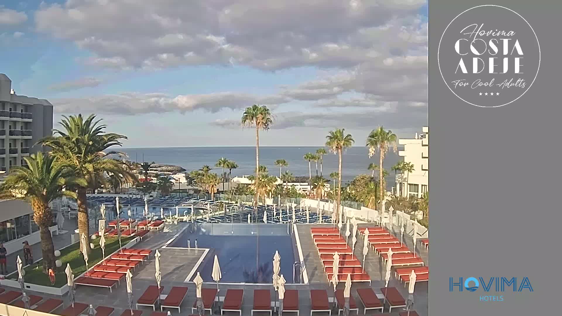 An outdoor pool area with many lounge chairs and umbrellas is situated next to a hotel, overlooking a calm ocean under a partly cloudy sky.