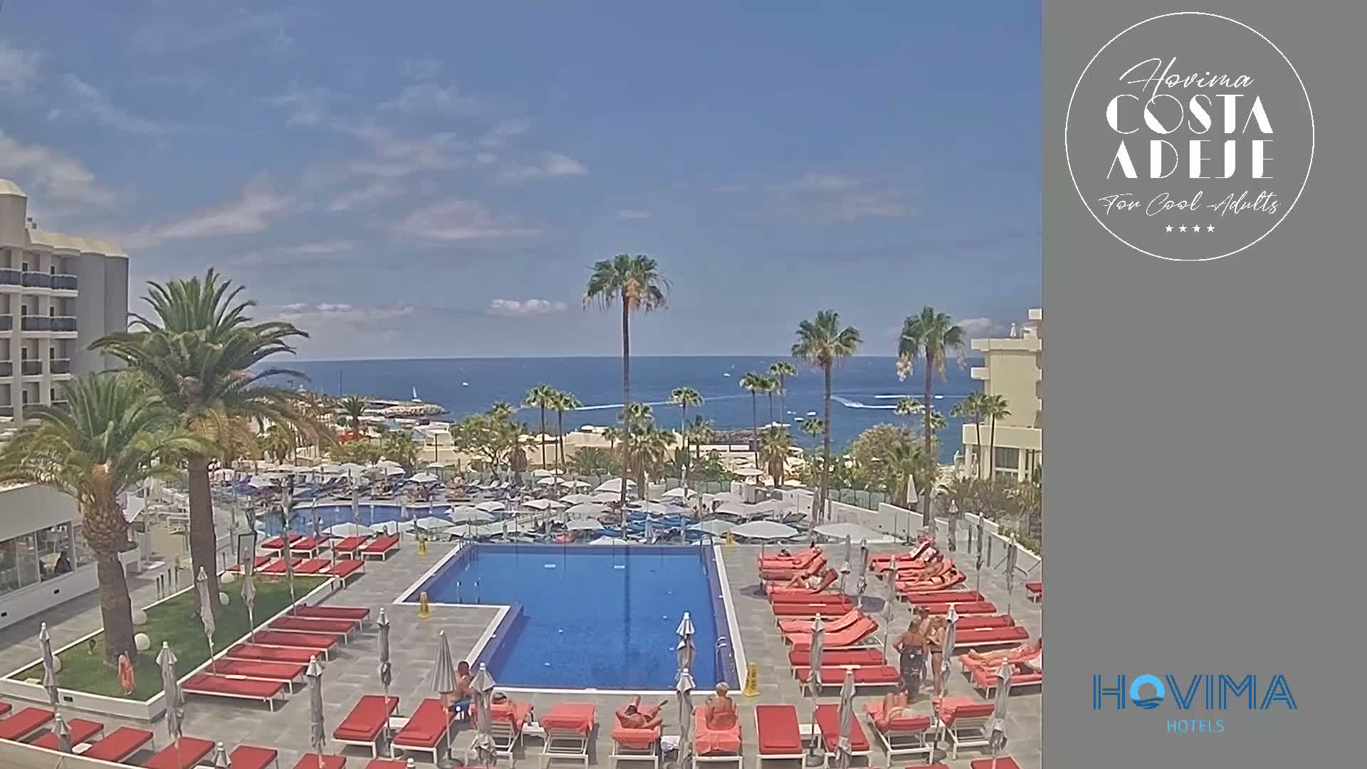 A resort pool area with red lounge chairs surrounds a rectangular pool, and palm trees and a calm ocean are visible in the background on a sunny day.