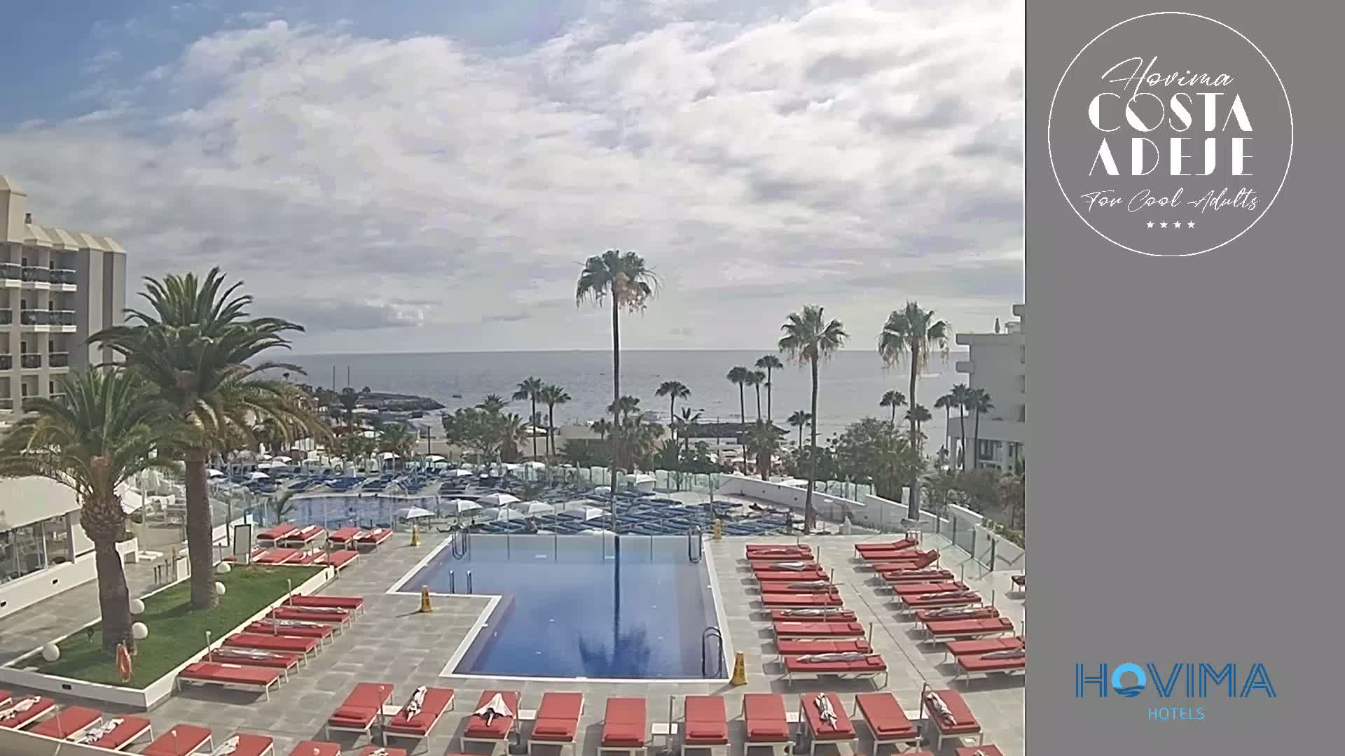 A resort pool area with red lounge chairs surrounds a rectangular swimming pool, overlooking a calm ocean under a partly cloudy sky.