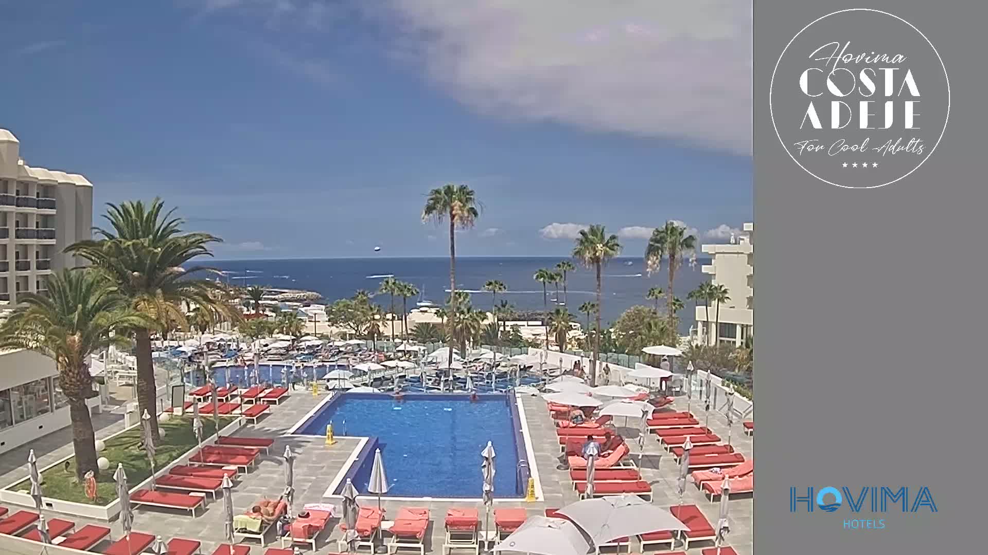 A resort pool area with red lounge chairs and palm trees surrounds a large swimming pool, overlooking a calm ocean under a mostly sunny sky.