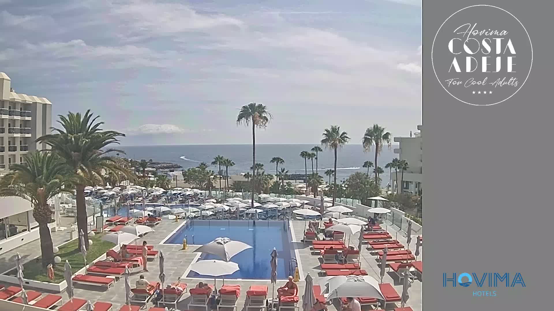 A resort pool area with many red lounge chairs and white umbrellas surrounds a blue swimming pool, overlooking a calm ocean on a sunny day.