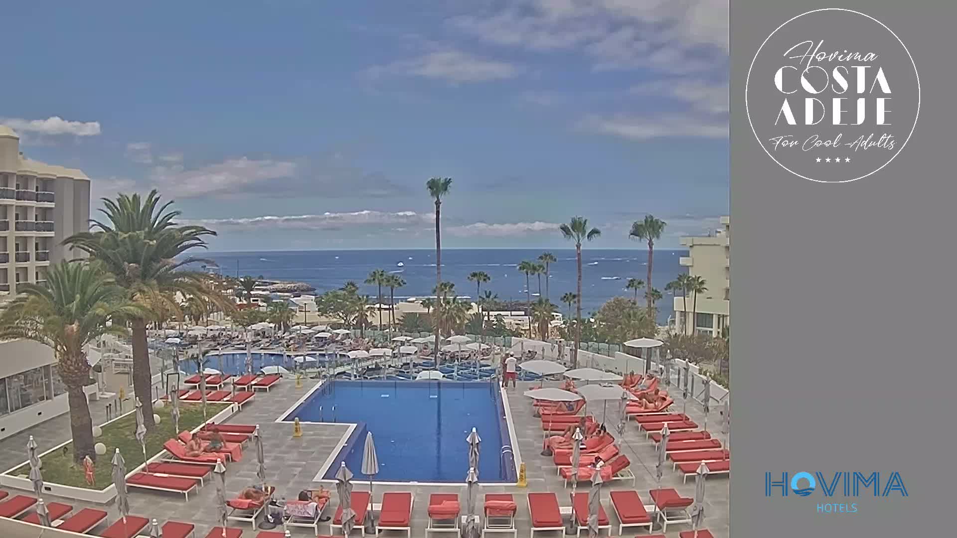 A resort pool area with red lounge chairs surrounds a large swimming pool, overlooking a calm ocean on a sunny day.