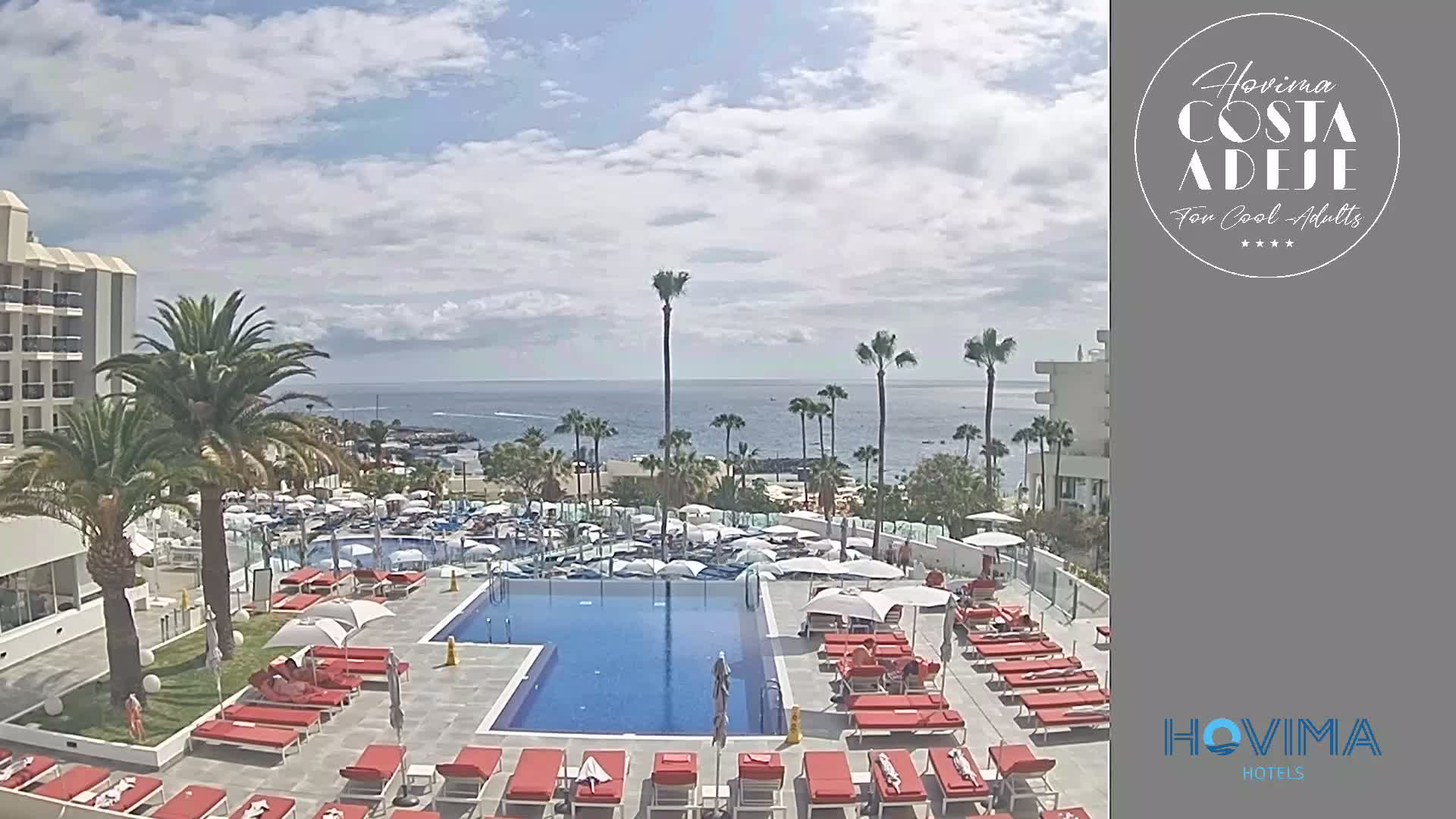 A resort pool area with red lounge chairs surrounds a swimming pool, overlooking a calm ocean under a partly cloudy sky.