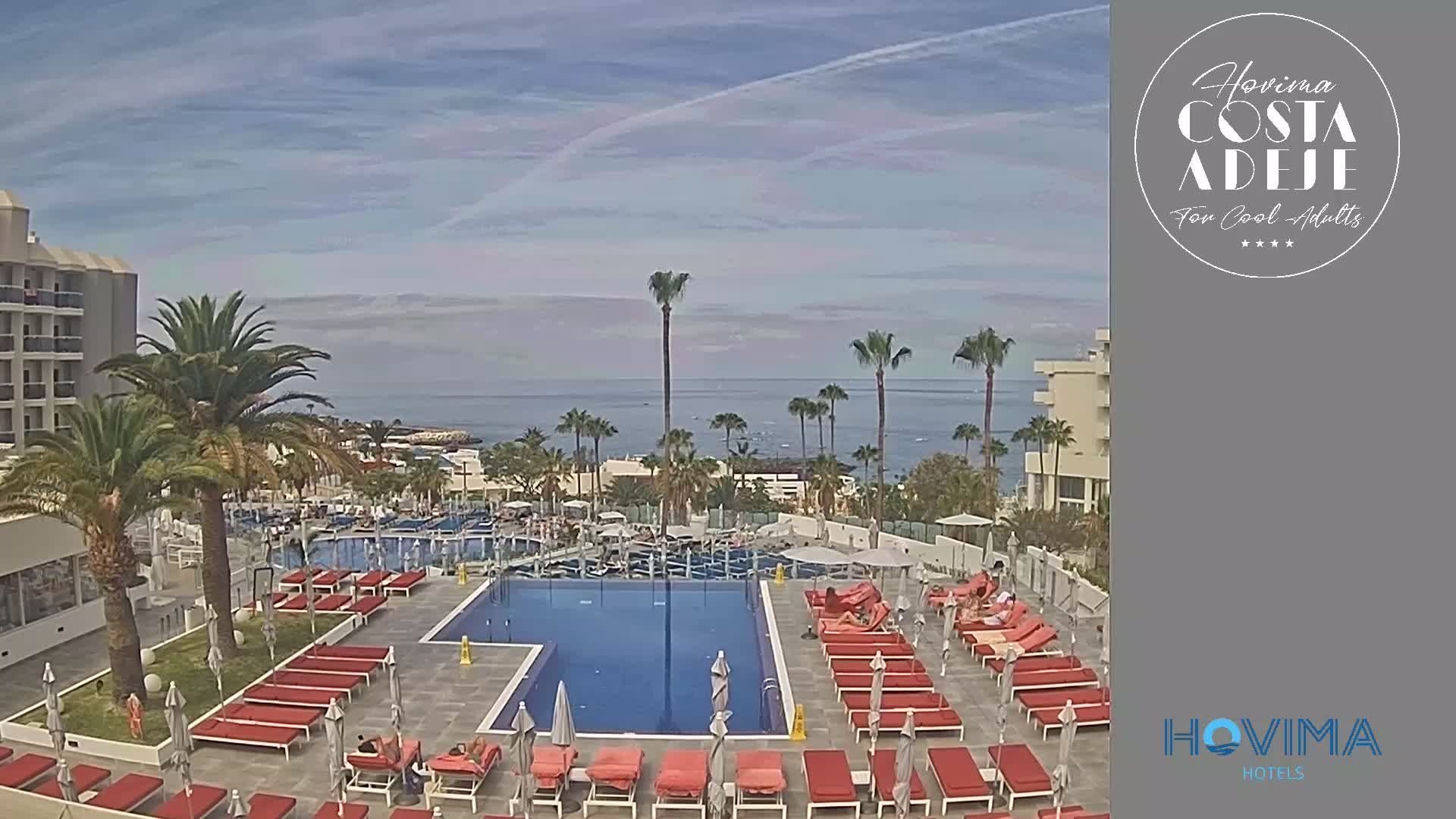A resort pool area with red lounge chairs surrounding a large swimming pool overlooks a calm ocean under a mostly clear sky.