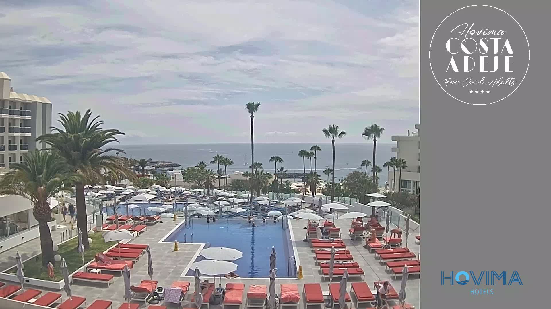 An outdoor pool area with red lounge chairs and umbrellas surrounding a large swimming pool, palm trees, and a hotel building overlooking a calm ocean under a partly cloudy sky.