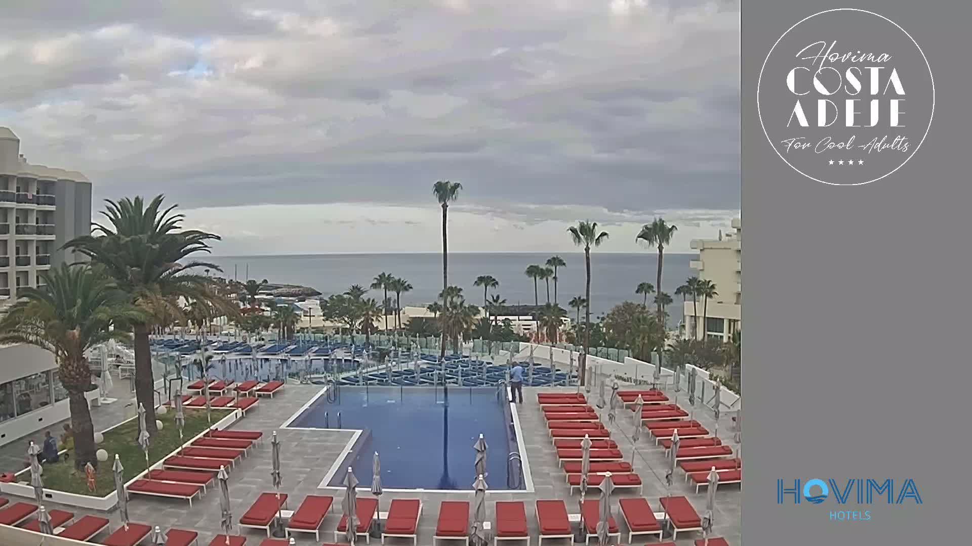 An outdoor swimming pool area with red lounge chairs surrounds a rectangular pool, overlooking a calm ocean under a cloudy sky.