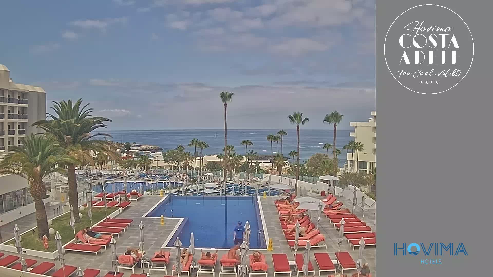 A resort pool area with red lounge chairs surrounds a large swimming pool, overlooking a calm ocean under a mostly sunny sky.