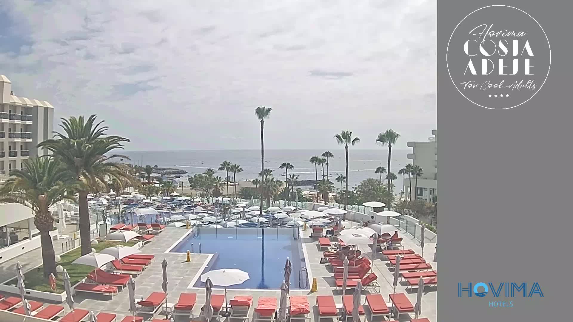 A hotel pool area with red lounge chairs and umbrellas overlooks a beach and calm ocean under a partly cloudy sky.