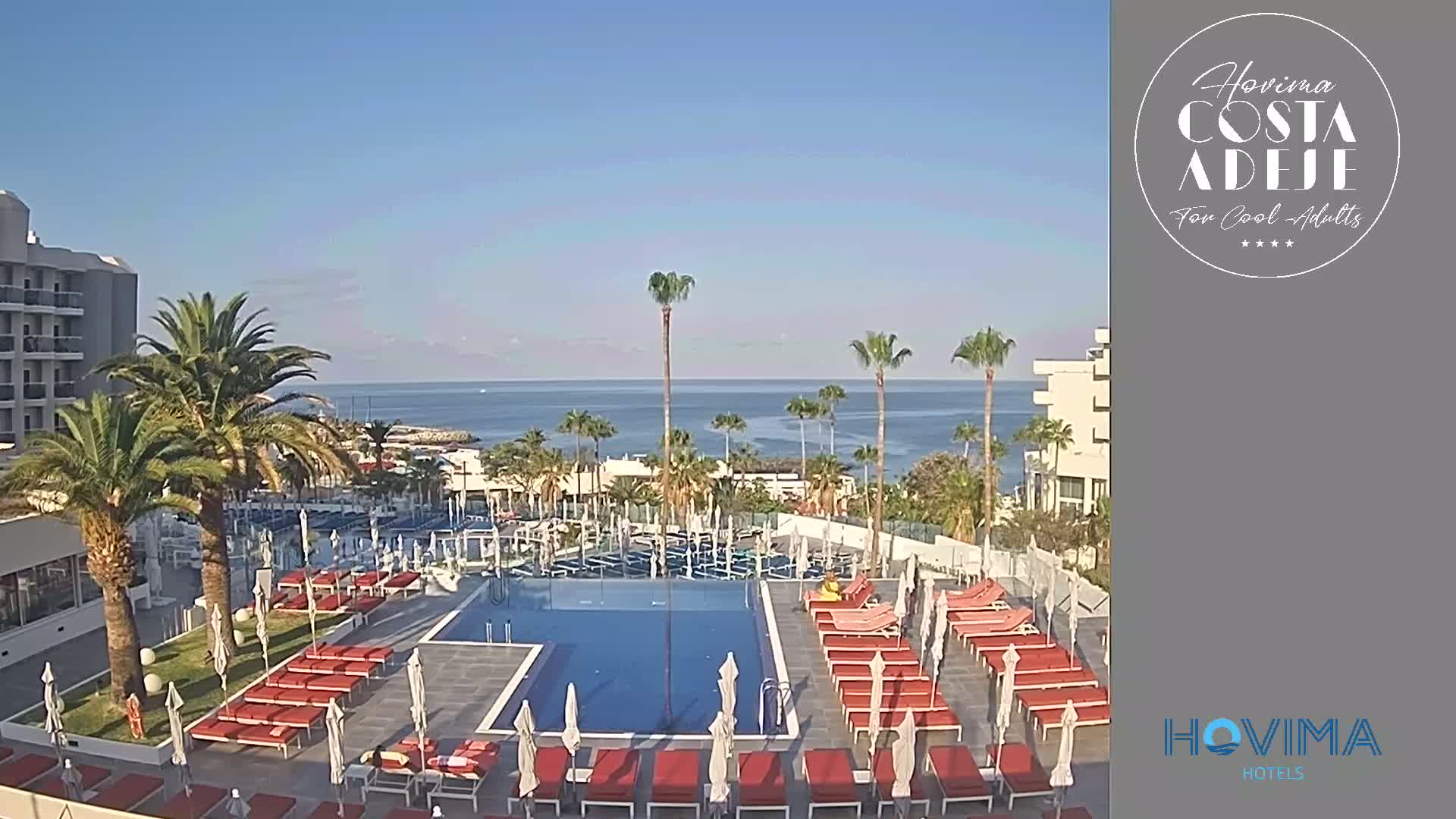 A hotel pool area with red lounge chairs and umbrellas overlooks a calm ocean under a clear blue sky.