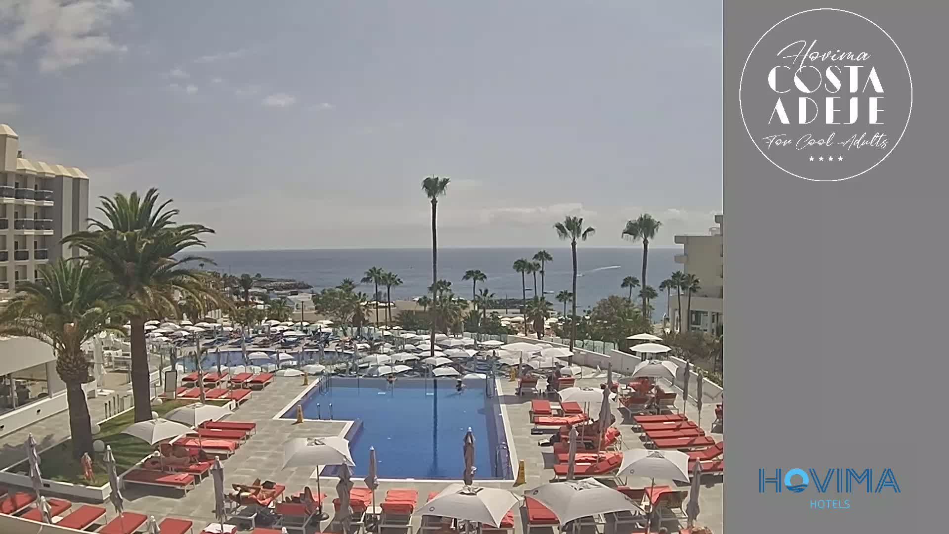 A resort pool area with red lounge chairs and umbrellas overlooks a calm ocean under a partly cloudy sky.