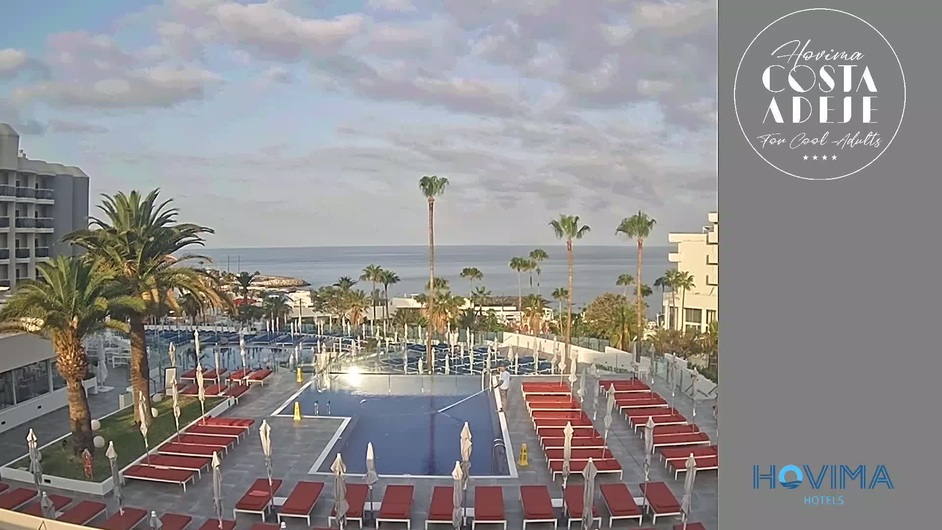 An outdoor swimming pool area with red lounge chairs and umbrellas, palm trees, and a building overlooks a calm ocean under a partly cloudy sky.