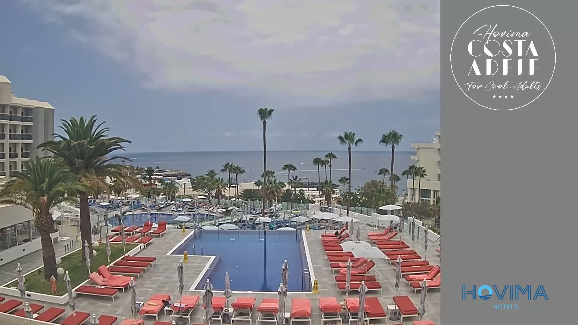 An outdoor pool area with red lounge chairs and umbrellas overlooks a calm ocean under a partly cloudy sky.