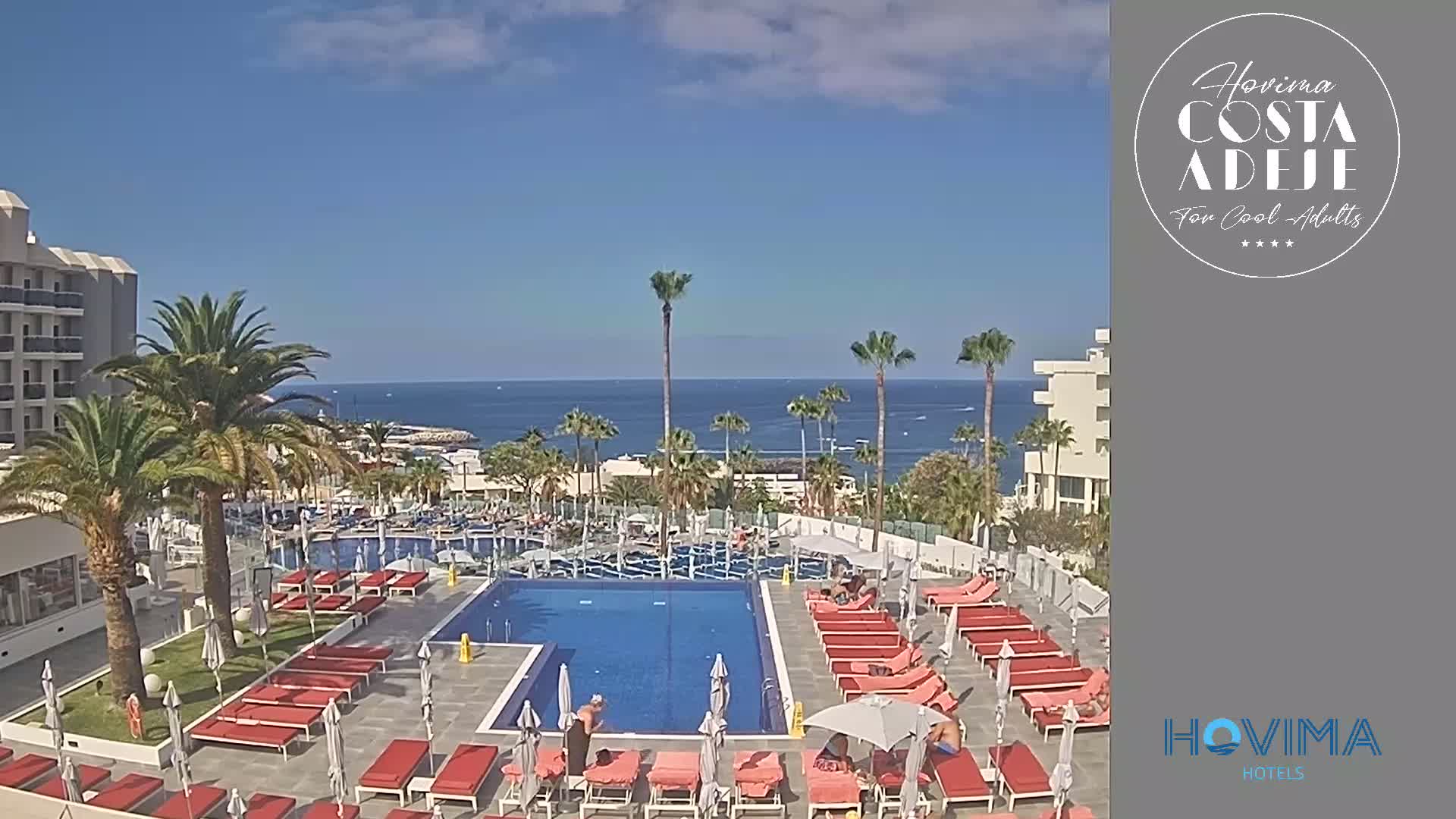 A resort pool area with red lounge chairs and umbrellas is situated next to a building overlooking a calm ocean on a sunny day.