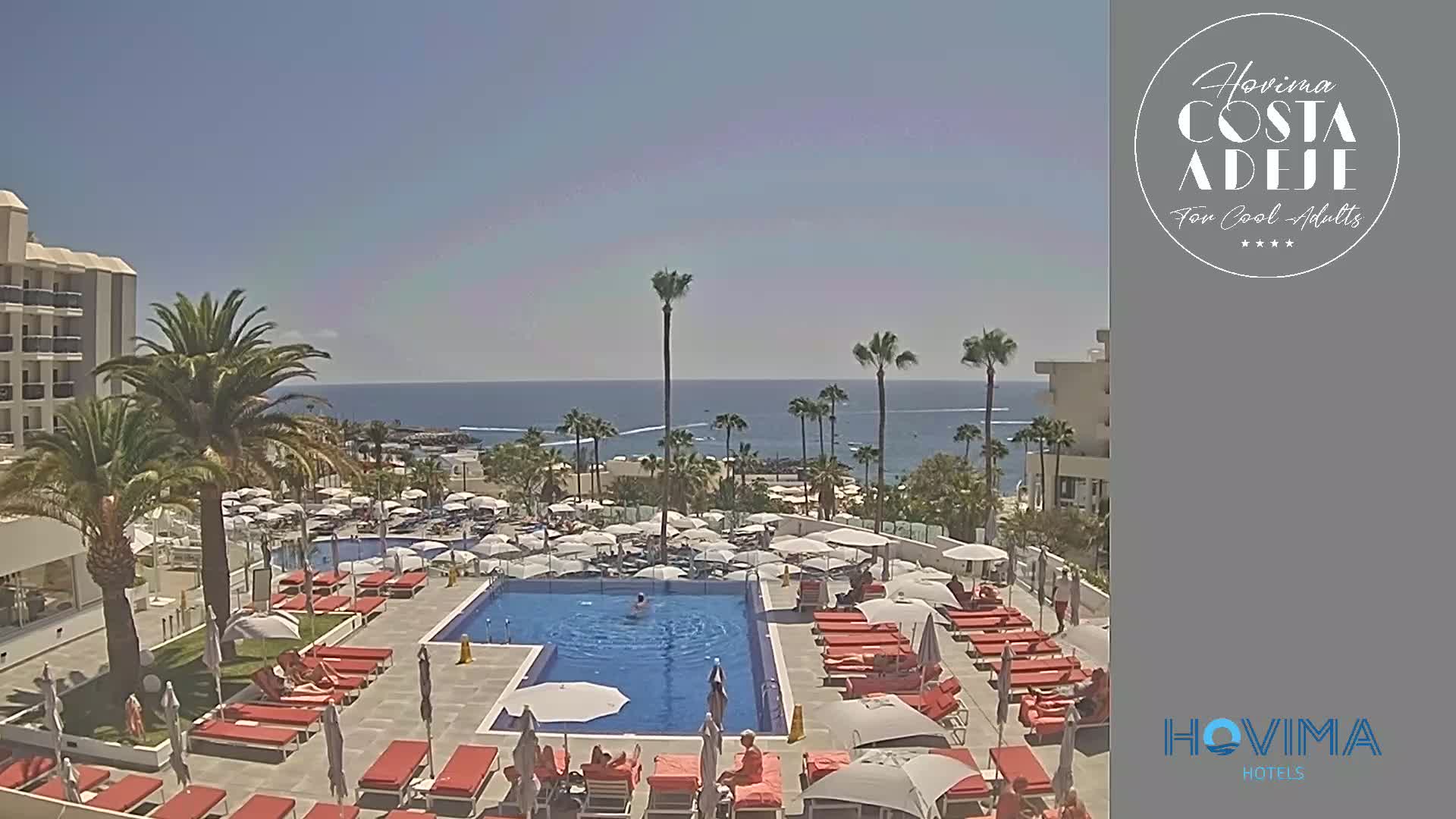 A resort pool area with red lounge chairs and numerous umbrellas surrounds a rectangular pool, overlooking a calm ocean under a clear blue sky.