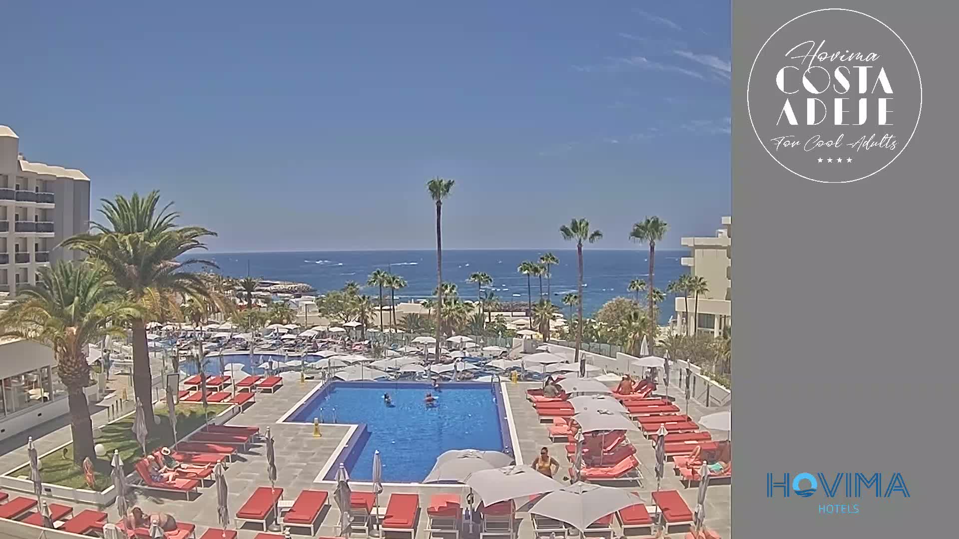 A resort pool area with red lounge chairs and umbrellas surrounds a large swimming pool, overlooking a calm ocean on a sunny day.