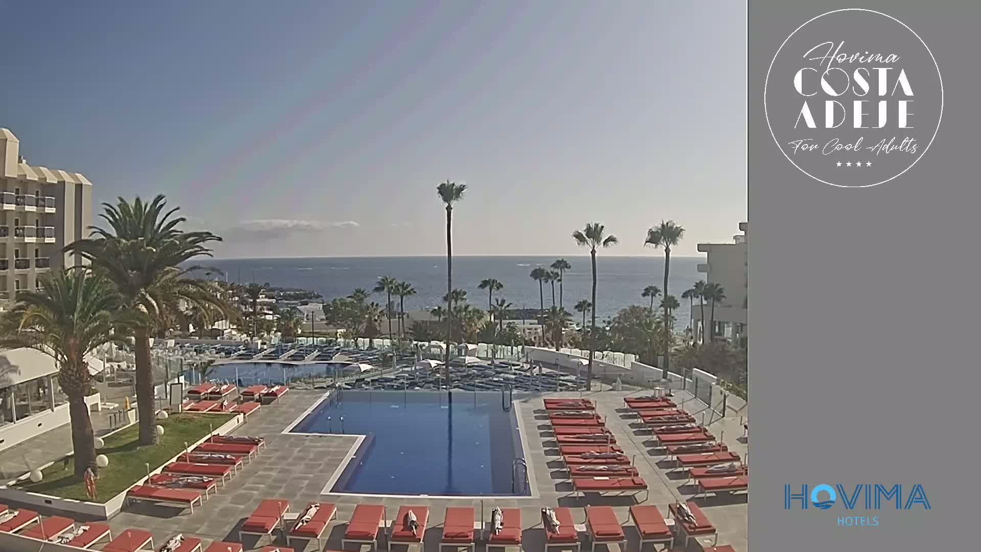 A resort pool area with red lounge chairs surrounds a large rectangular pool, overlooking a calm ocean under a clear blue sky.