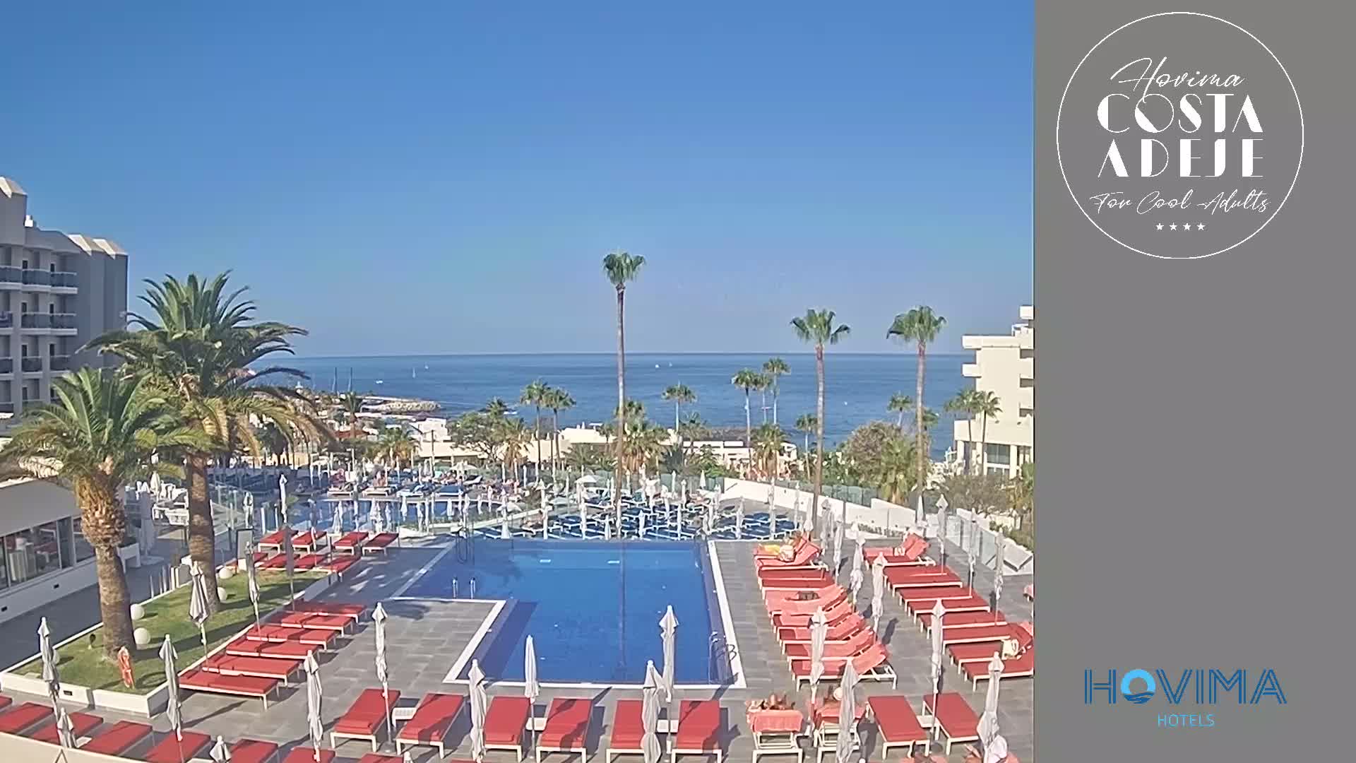 A resort pool area with red lounge chairs and umbrellas surrounds a rectangular pool, overlooking a calm ocean on a sunny day.