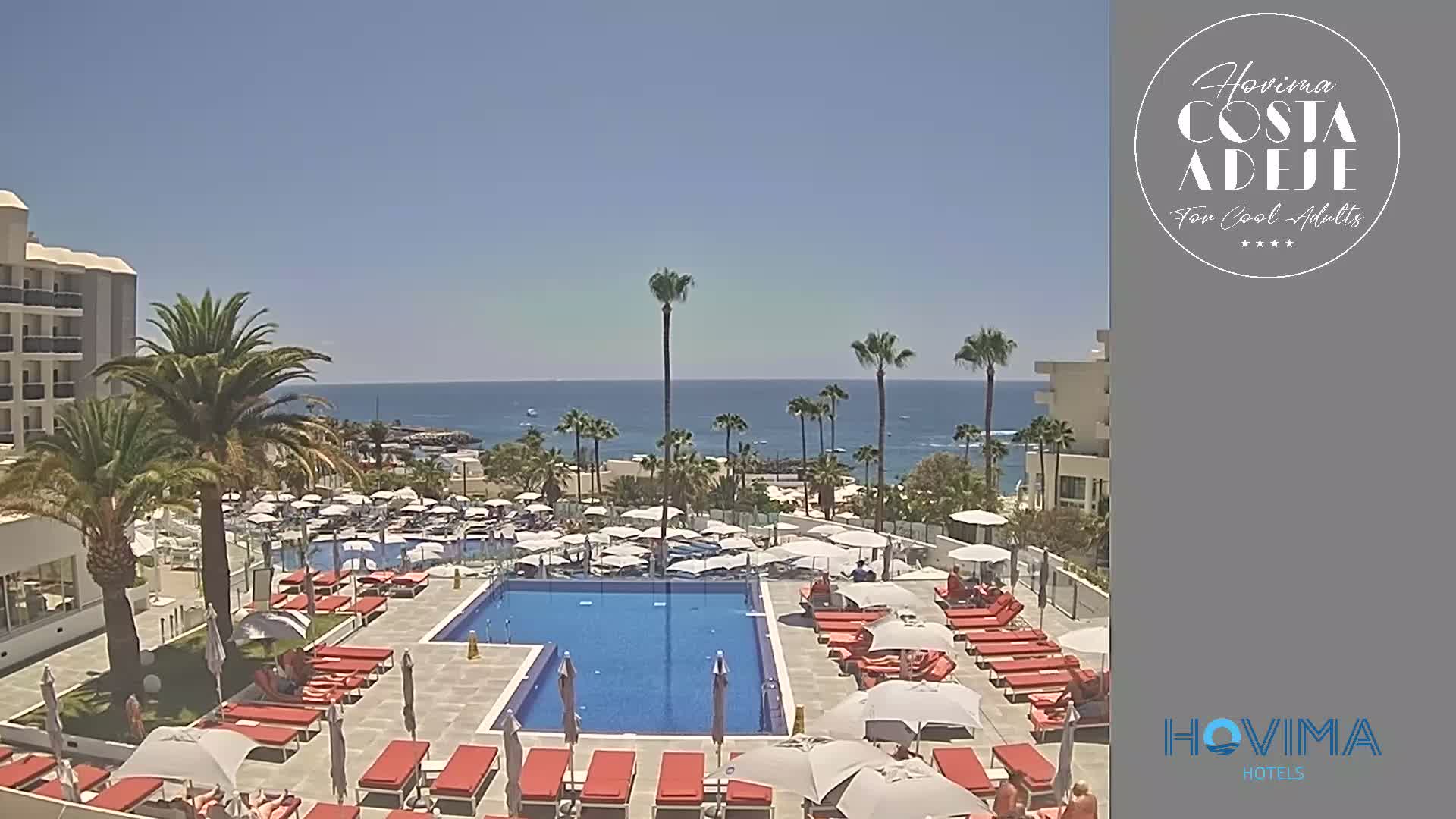 A resort pool area with red lounge chairs and umbrellas surrounds a large swimming pool, overlooking a calm ocean on a sunny day.