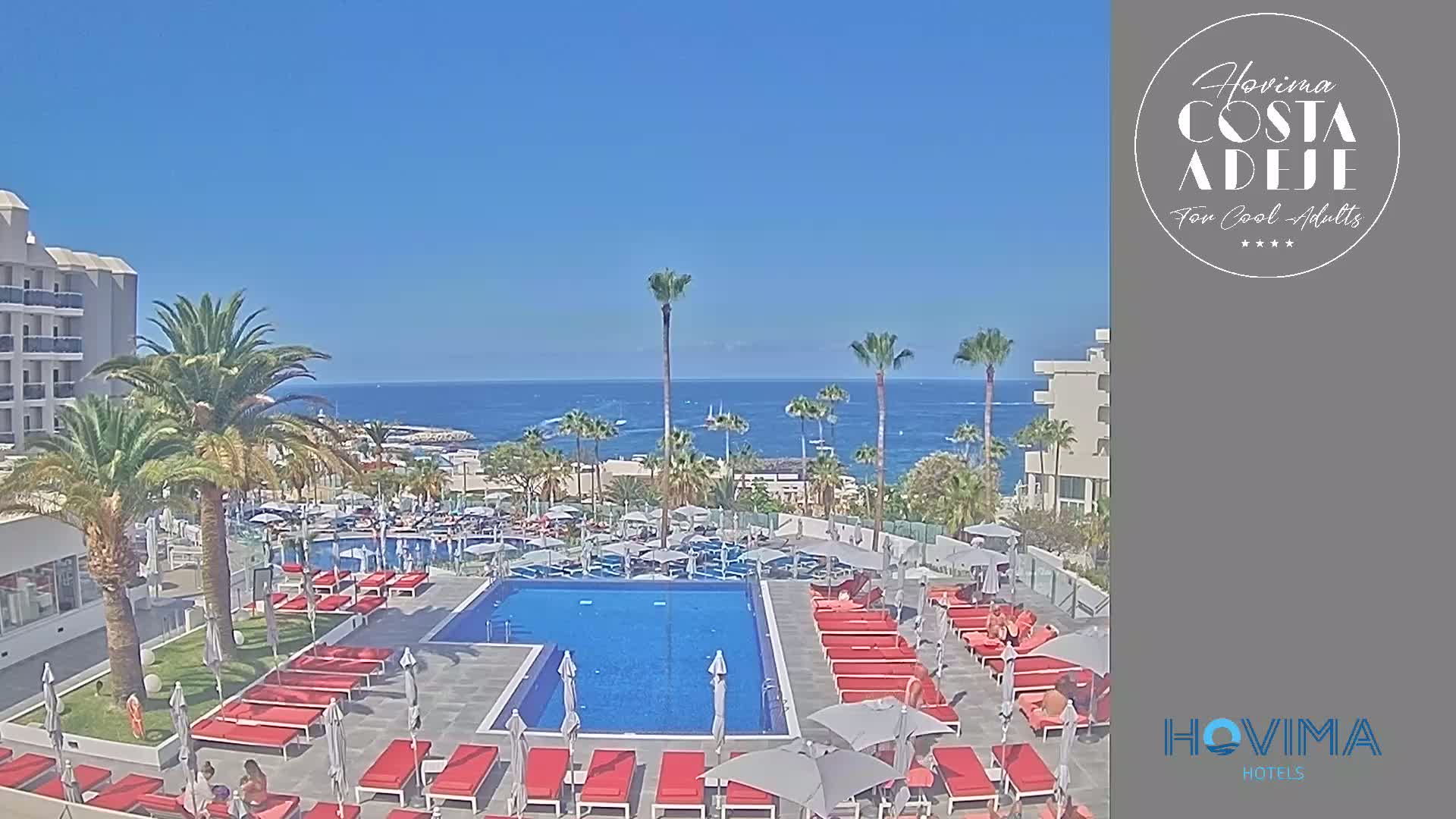 A resort pool area with red lounge chairs and umbrellas surrounds a rectangular pool, all situated near a calm ocean under a clear blue sky.
