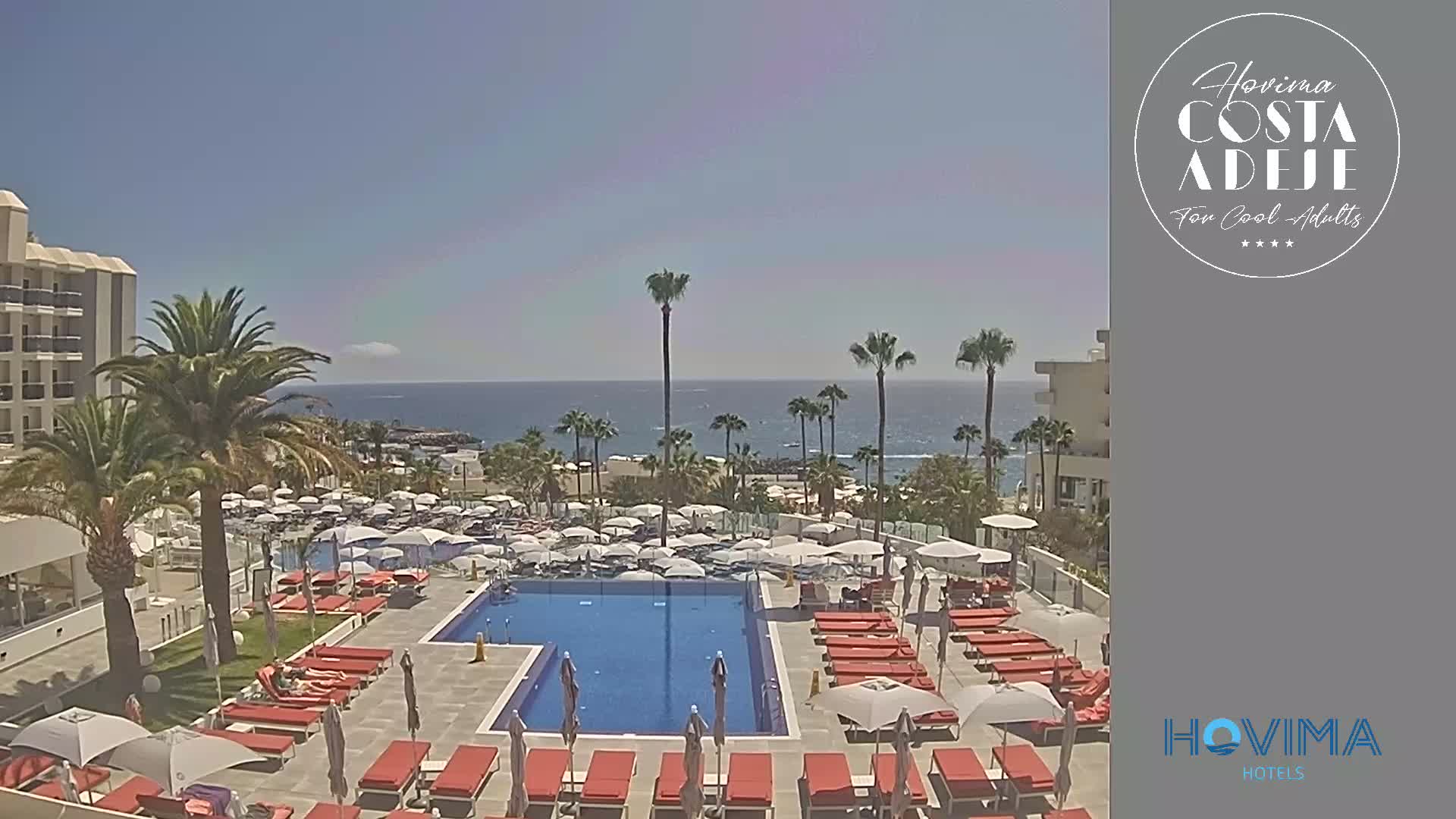 A resort pool area with numerous red lounge chairs and white umbrellas surrounds a large, rectangular pool, all overlooking a calm ocean under a clear, sunny sky.