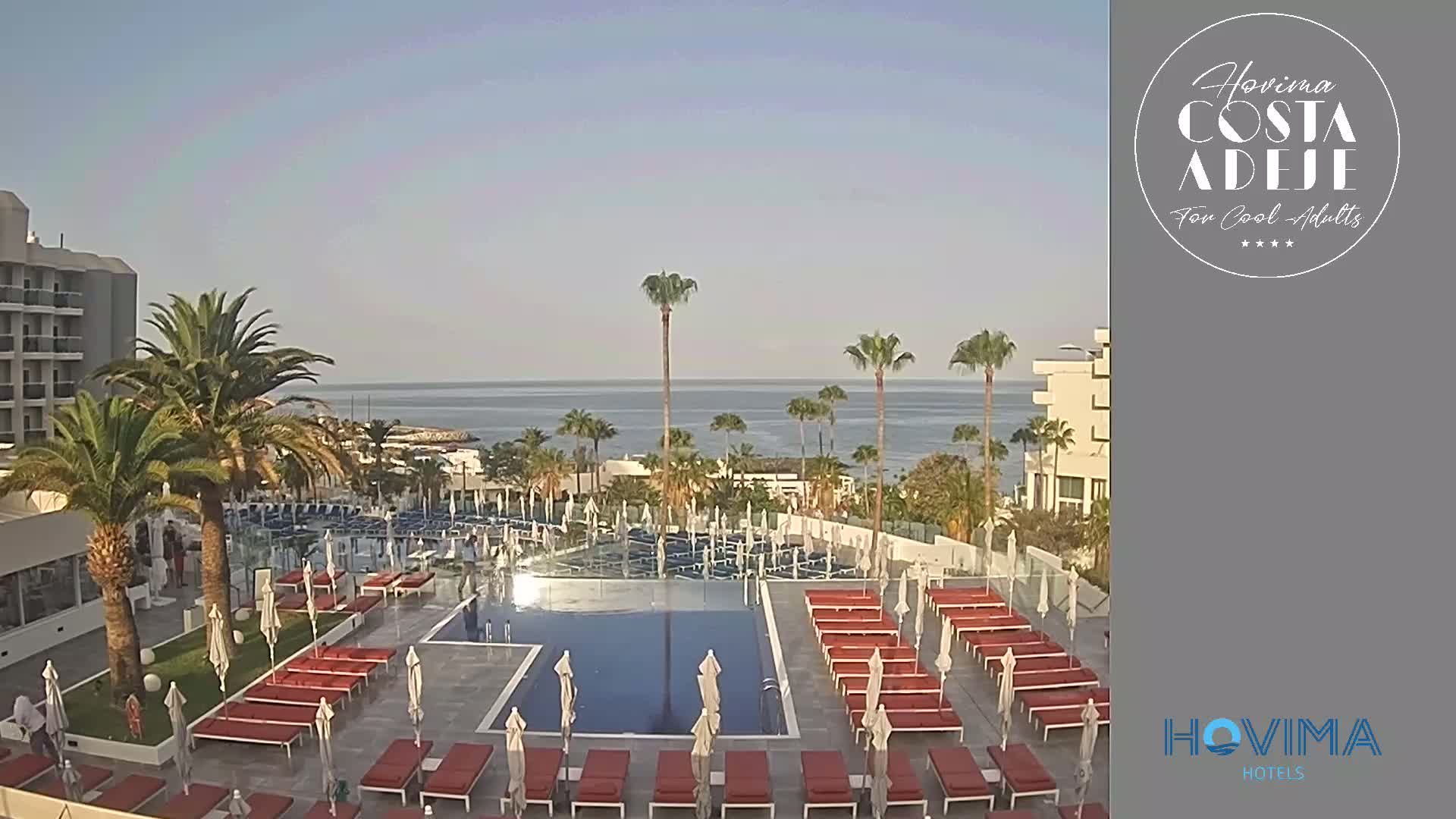 A resort pool area with many lounge chairs and umbrellas is situated next to a building overlooking a calm ocean under a clear sky.