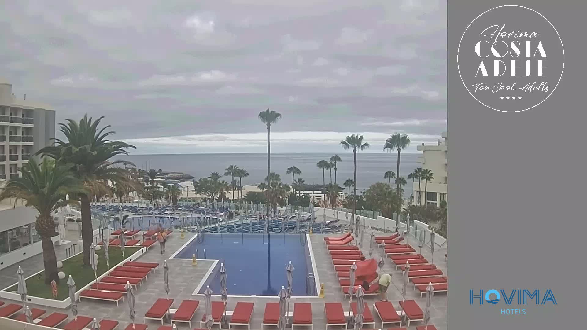 An overcast day reveals a resort pool area with red lounge chairs surrounding a large rectangular pool, overlooking a calm ocean and palm trees.