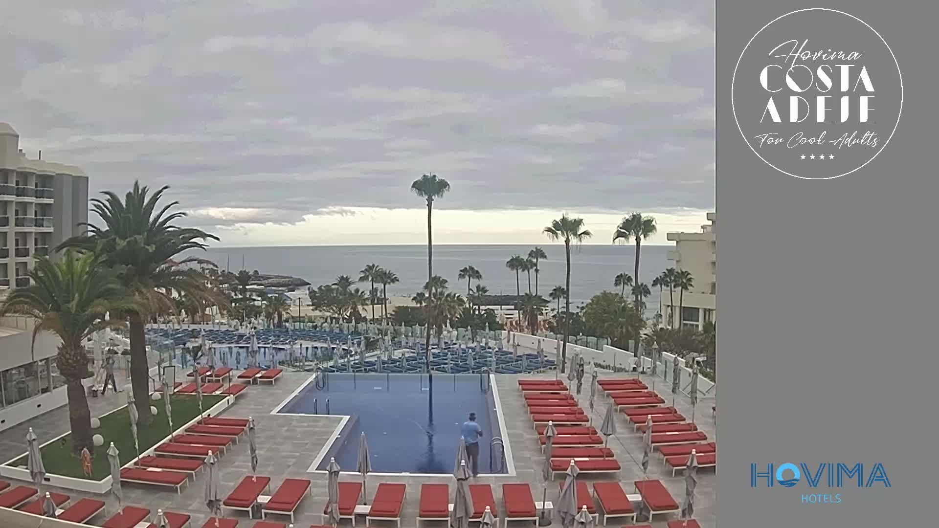 A resort pool area with red lounge chairs and umbrellas overlooks a calm ocean under an overcast sky.