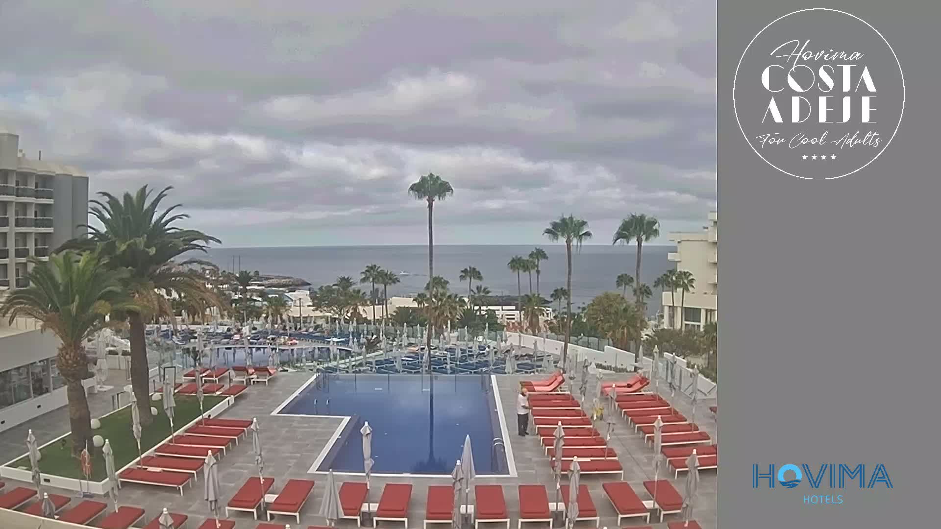 An outdoor pool area with red lounge chairs and palm trees overlooks a calm ocean under a cloudy sky.