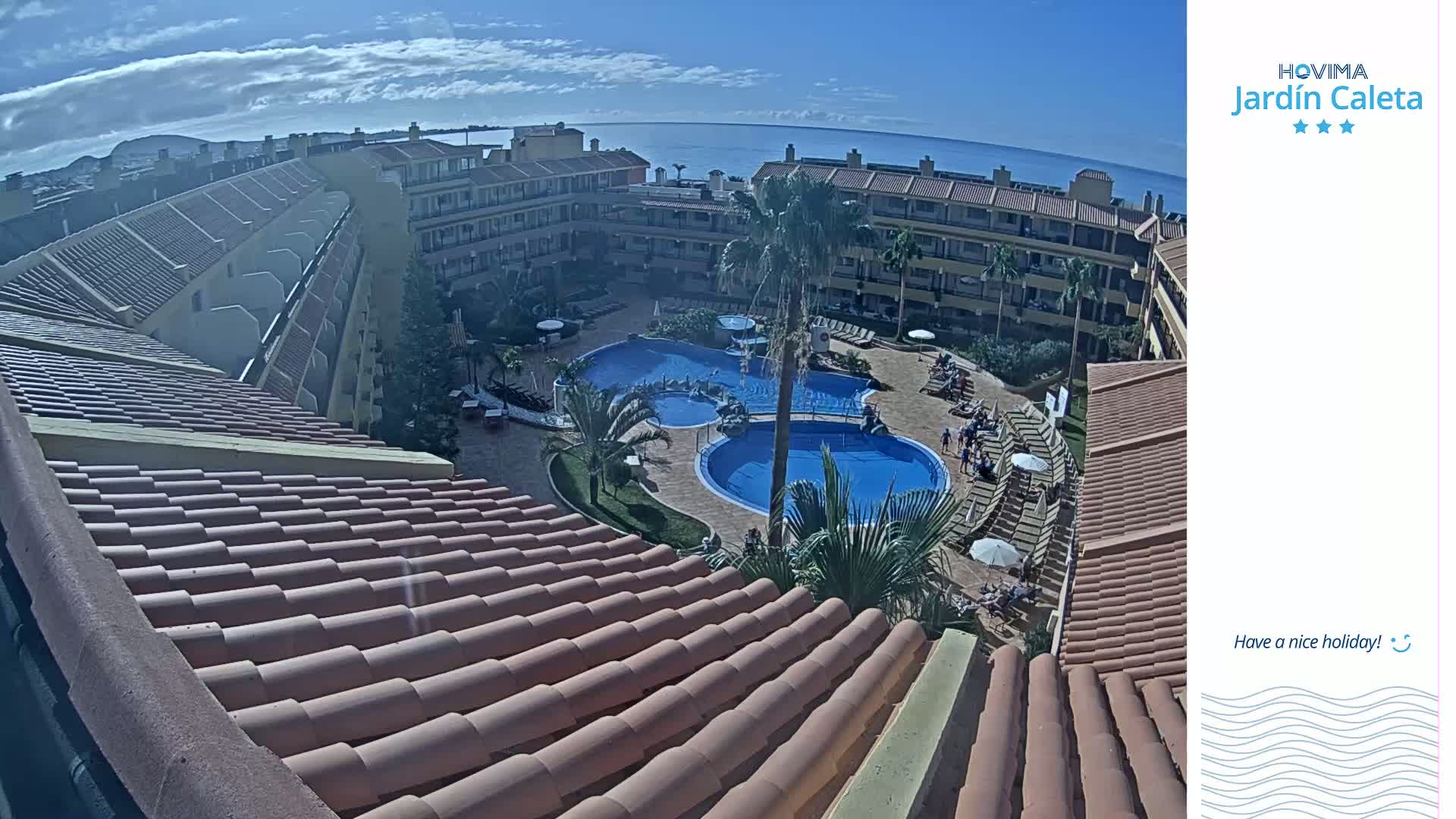 A high-angle view of a resort courtyard with two pools surrounded by buildings under a mostly clear, sunny sky.