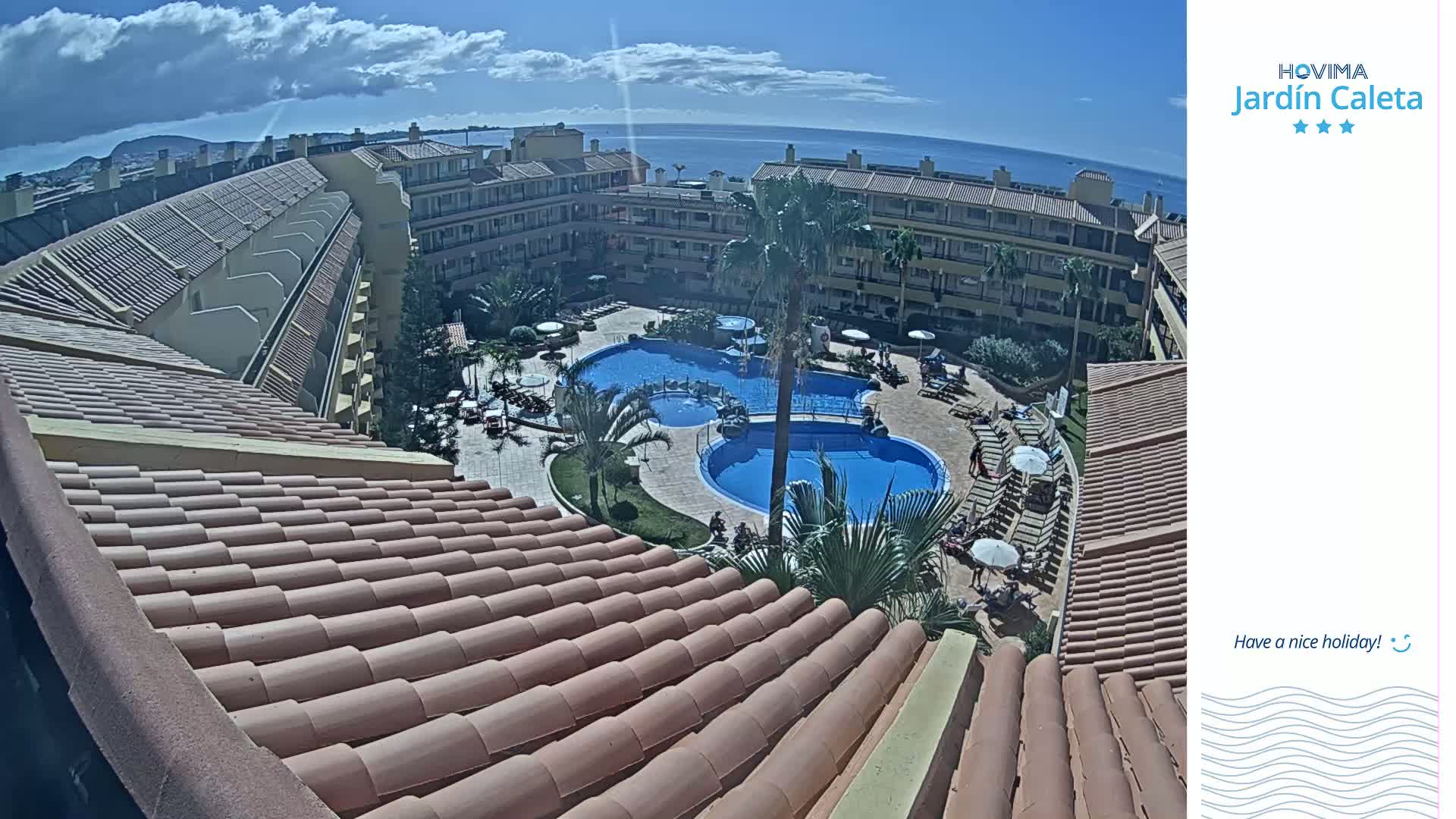 A tiled rooftop overlooks a courtyard with two swimming pools, surrounded by a building and palm trees, under a partly cloudy sky with a view of the ocean in the distance.