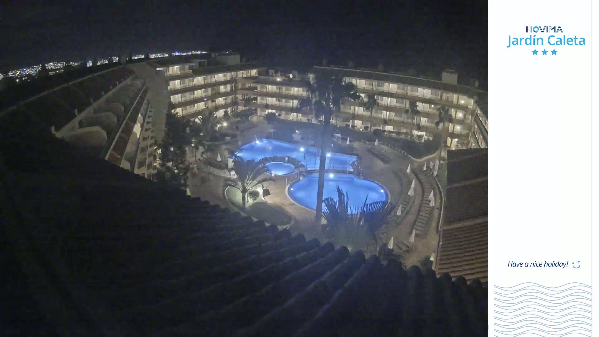 A nighttime view from a rooftop shows a multi-story building complex surrounding a lit swimming pool area.