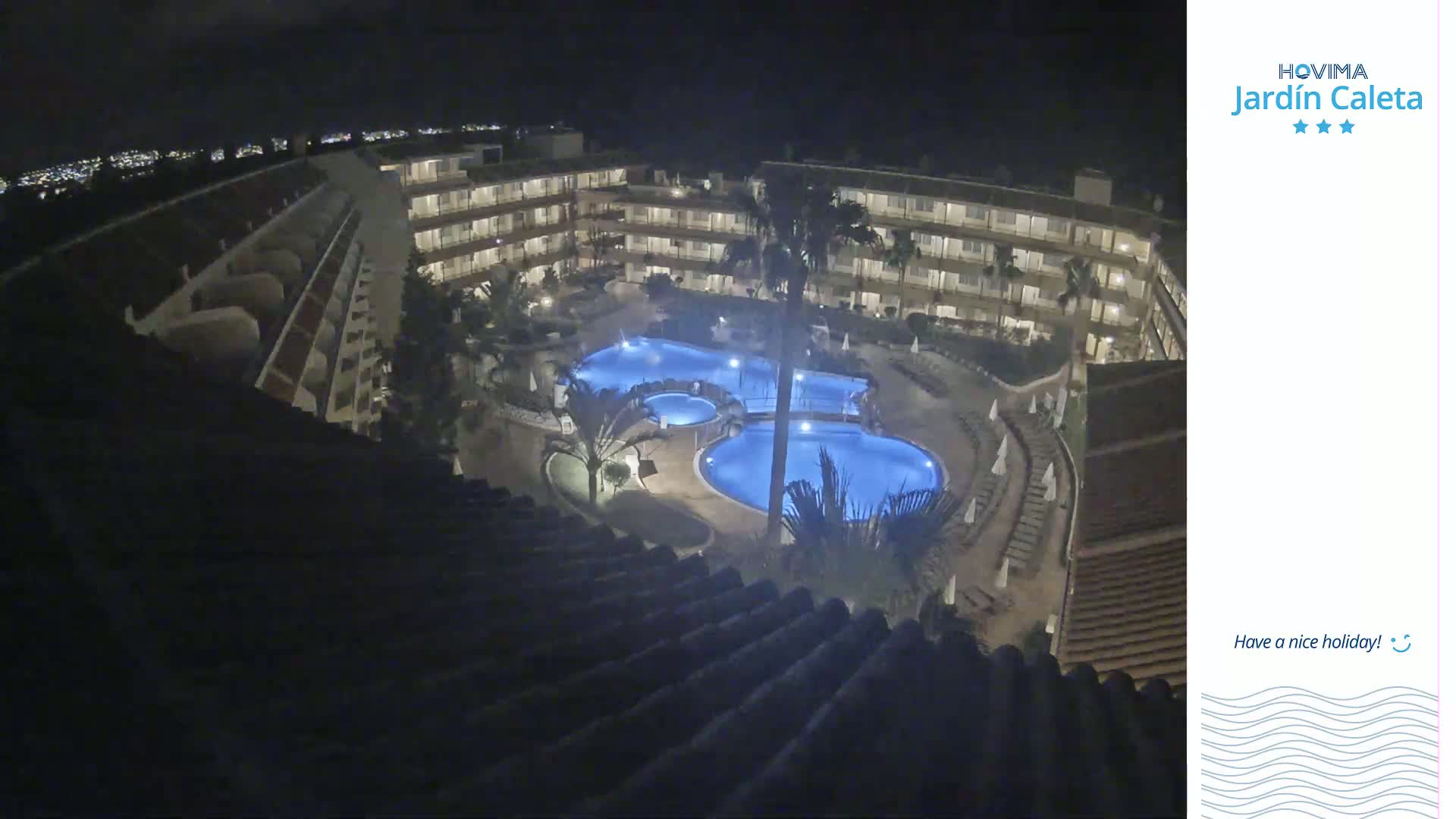 A nighttime aerial view shows a resort complex with illuminated swimming pools surrounded by buildings and palm trees.