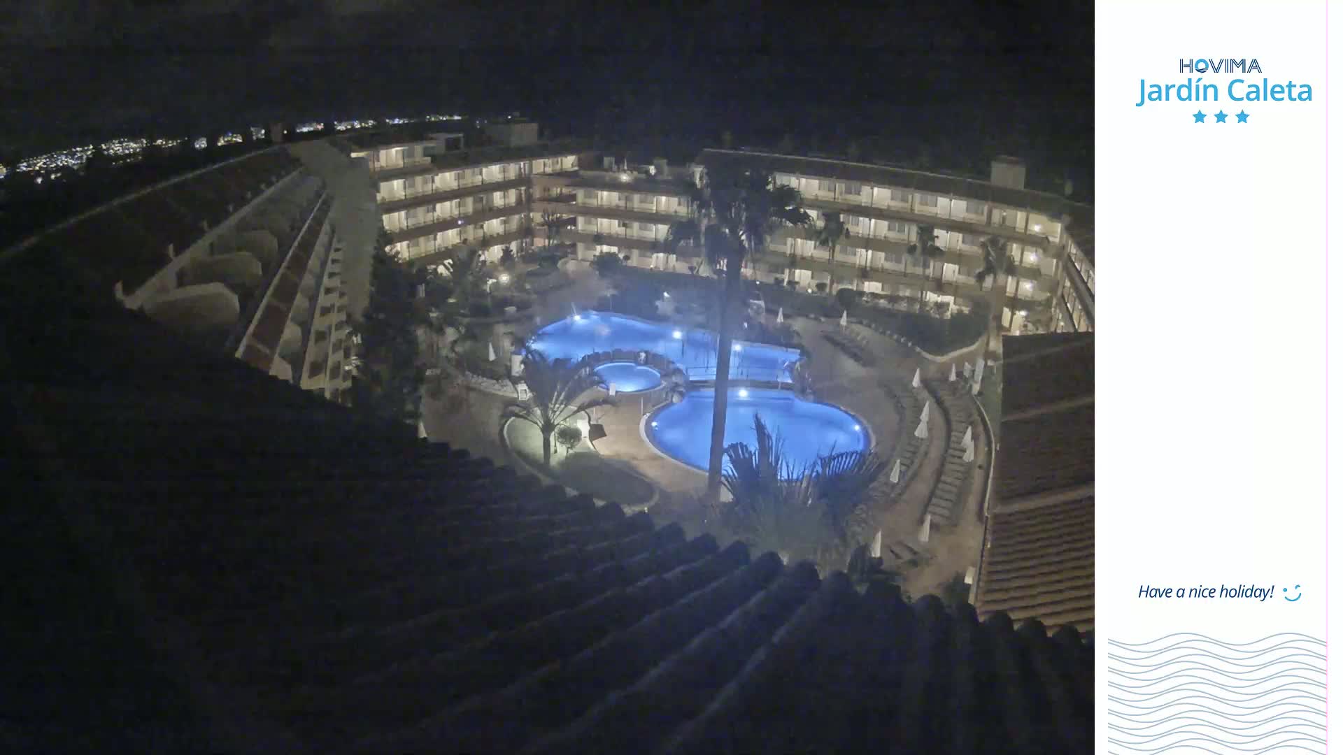 A nighttime, high-angle view of a resort complex with multiple illuminated swimming pools surrounded by buildings and palm trees.