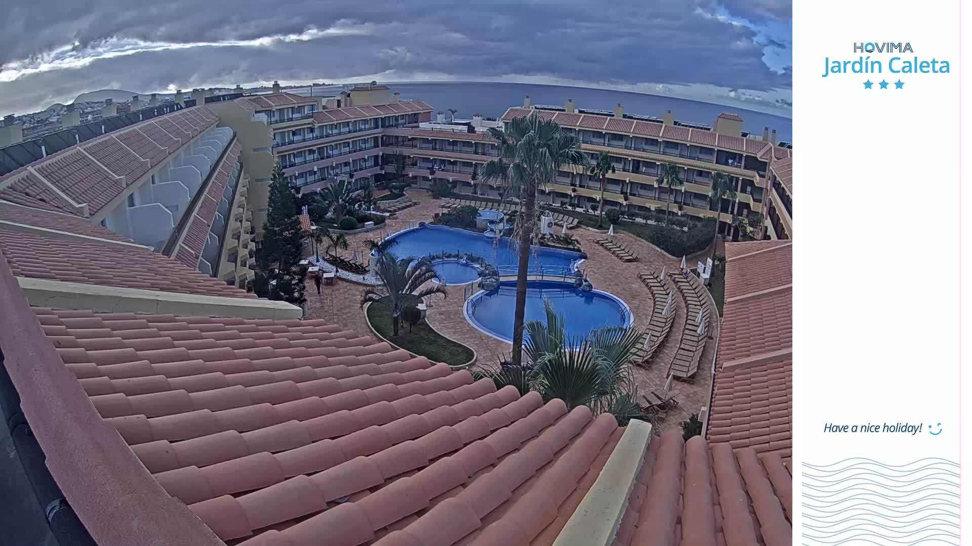 An overhead view shows a resort with two swimming pools surrounded by lounge chairs and palm trees, under an overcast sky.