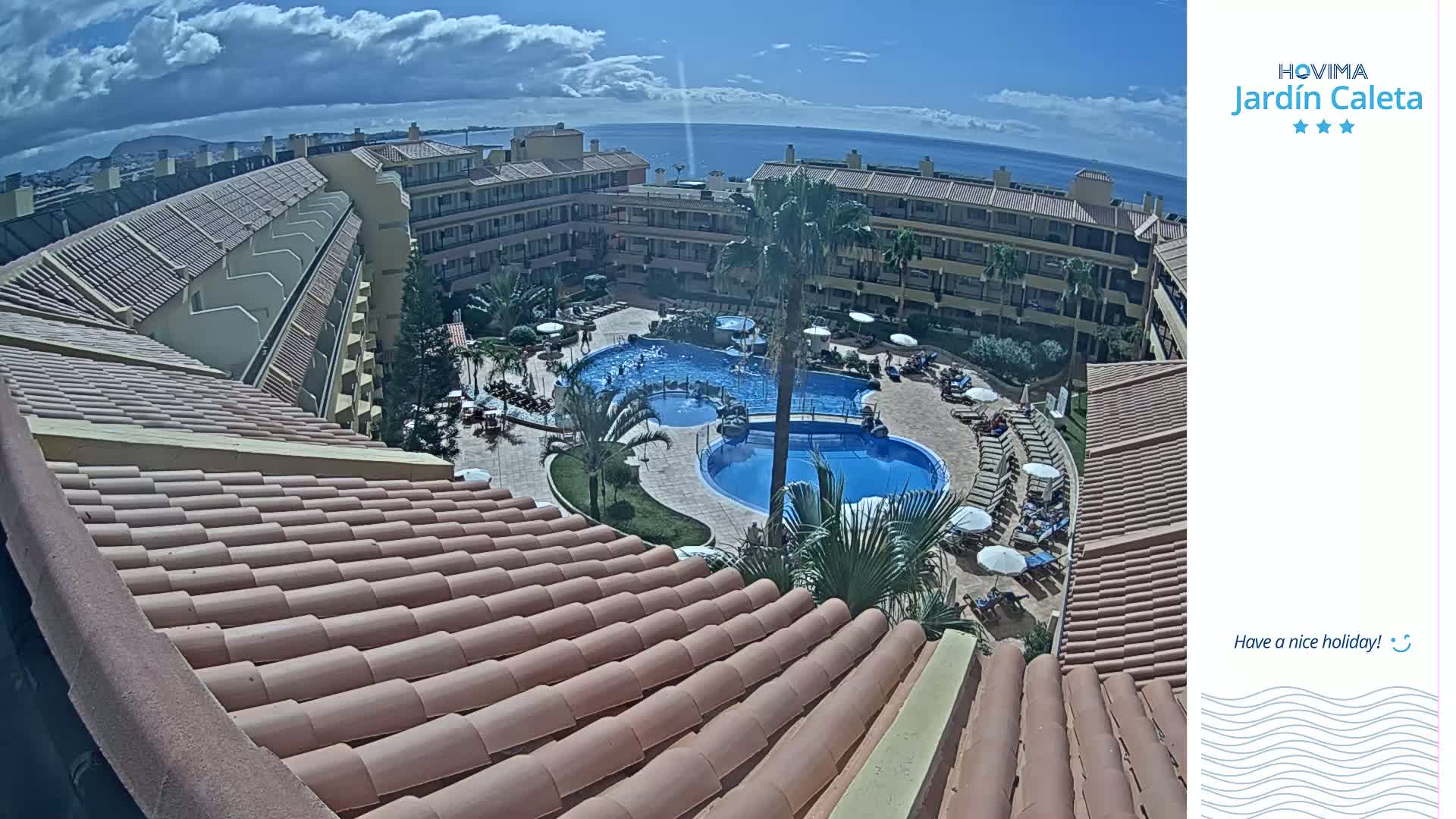 An aerial view shows a resort with multiple pools surrounded by buildings under a partly cloudy sky, overlooking a distant ocean.